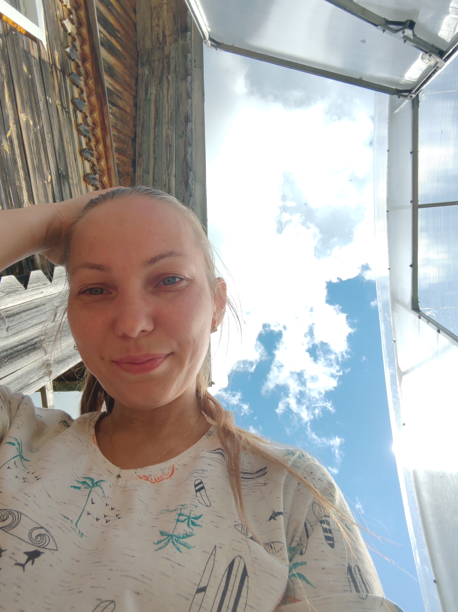 A young woman is captured in a close-up, low-angle selfie outdoors, with a wooden structure on the left and a translucent roof of a structure overhead. The background is dominated by a bright blue sky with scattered white clouds, suggesting a sunny day. The woman has fair skin, light-colored eyes, and her blonde hair is pulled back with one hand resting on her head. She is wearing a light-colored t-shirt with a pattern of palm trees, birds, and surfboards. The setting appears to be a rural or suburban area, possibly a dacha (Russian country house) given the wooden construction, and the mention of Чухлэм, Russia, suggests a specific location. The image has a casual, informal feel, typical of a personal photograph.