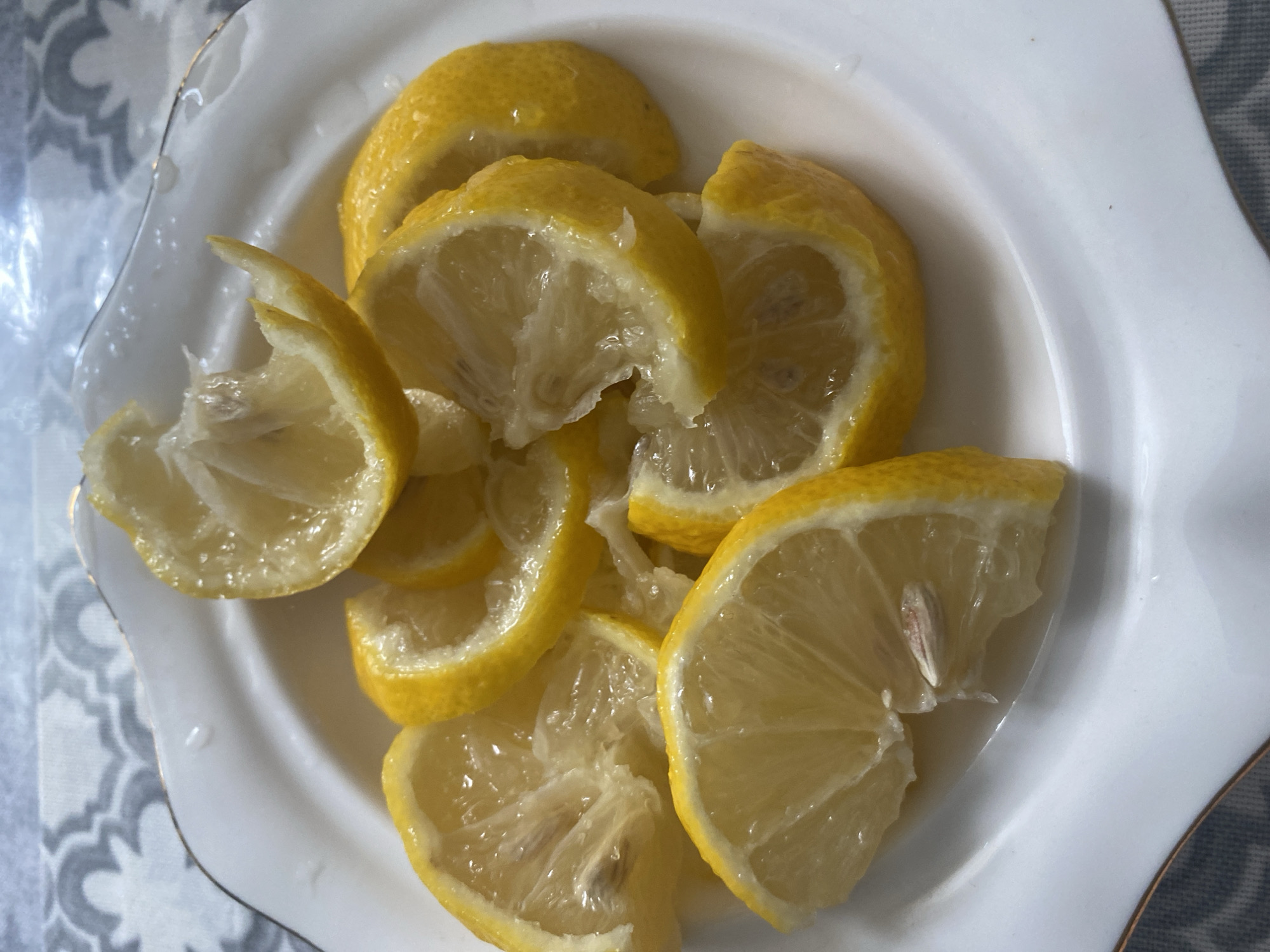 A close-up shot features a white, scalloped-edge plate filled with several bright yellow lemon slices. The plate has a thin, gold rim. The lemons are cut into various wedge shapes, some showing their juicy pulp and a few with visible seeds. The surface of the plate is slightly wet, indicating the juice from the lemons. The background is blurred, showing a grey and white patterned cloth. There is no discernible location in the city of Chontaul, Russia, visible in this image.