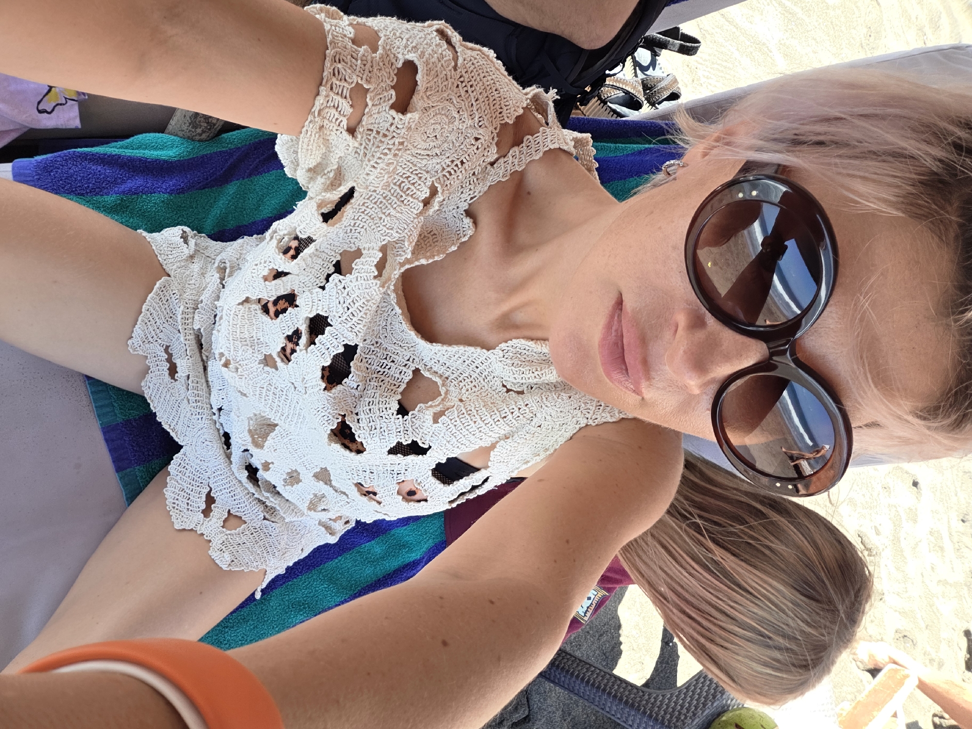 The original image is a selfie taken by a woman with short, light pink hair. She is wearing large, dark brown, round sunglasses and a cream-colored, crocheted top with cutouts. Underneath the top, a leopard-print bikini top is visible. The woman is sitting in a beach chair on a sandy beach, as indicated by the sand visible in the background and the reflection in her sunglasses showing a person walking by the water. She is using her left arm to take the photo. To her right, another person with blonde hair is seated, and their upper body is partially visible. A colorful striped beach towel is draped behind the woman. The lighting suggests it is daytime, likely sunny, given the bright beach environment. The location is identified as North Kuta, Indonesia.