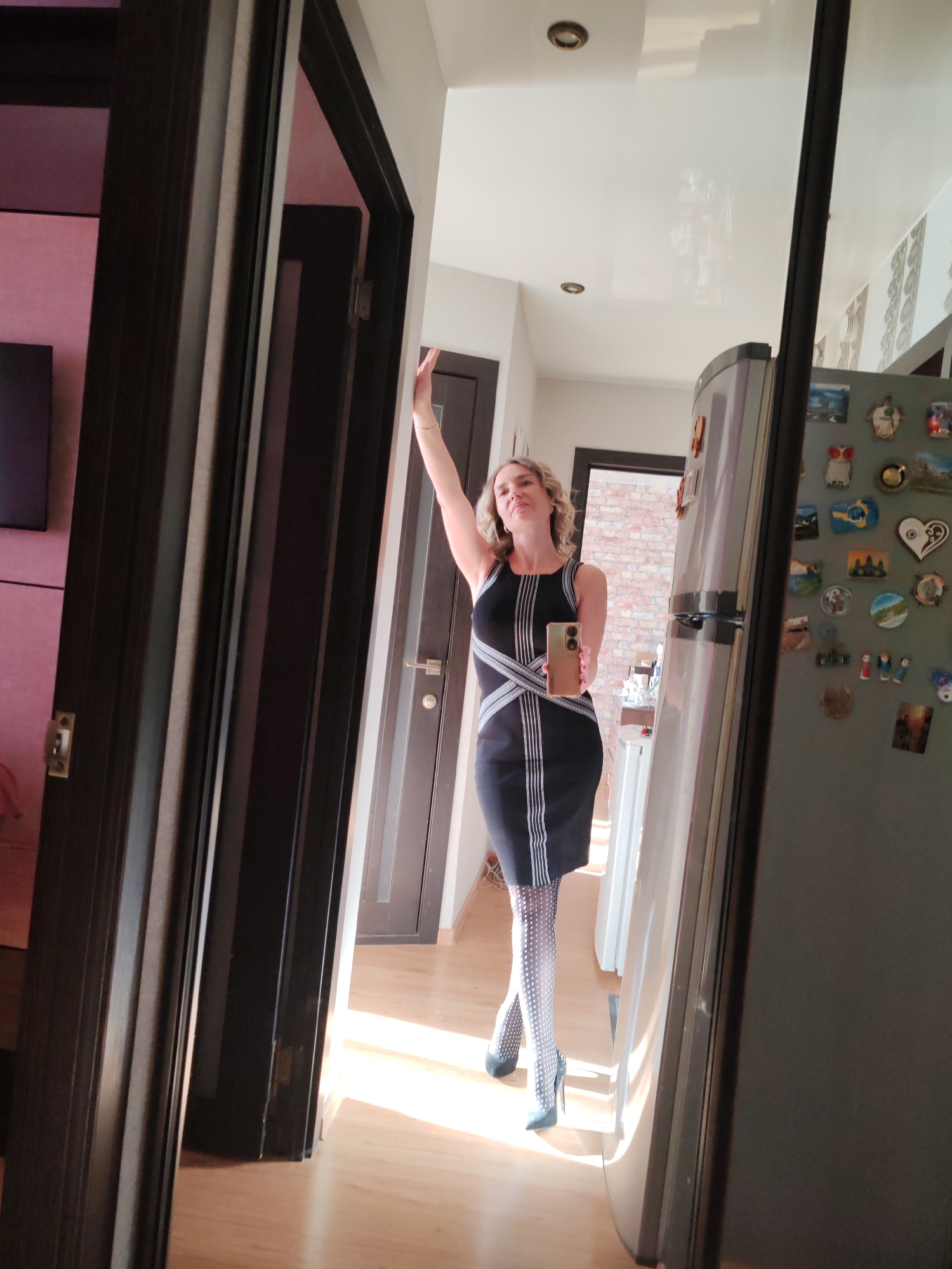 A woman in a black dress and polka-dot stockings poses for a selfie in front of a mirror. She is holding a phone in her right hand and has her left arm raised. The mirror reflects a kitchen interior with a refrigerator adorned with magnets. The room appears to be well-lit. Given the presence of Russian-style fridge magnets on the refrigerator, such as one featuring the bear and salmon symbol of Kamchatka Krai and another featuring a stylized volcano, it is likely that the location of the photo is in the city of Petropavlovsk-Kamchatsky, Russia or somewhere else in the Krai.