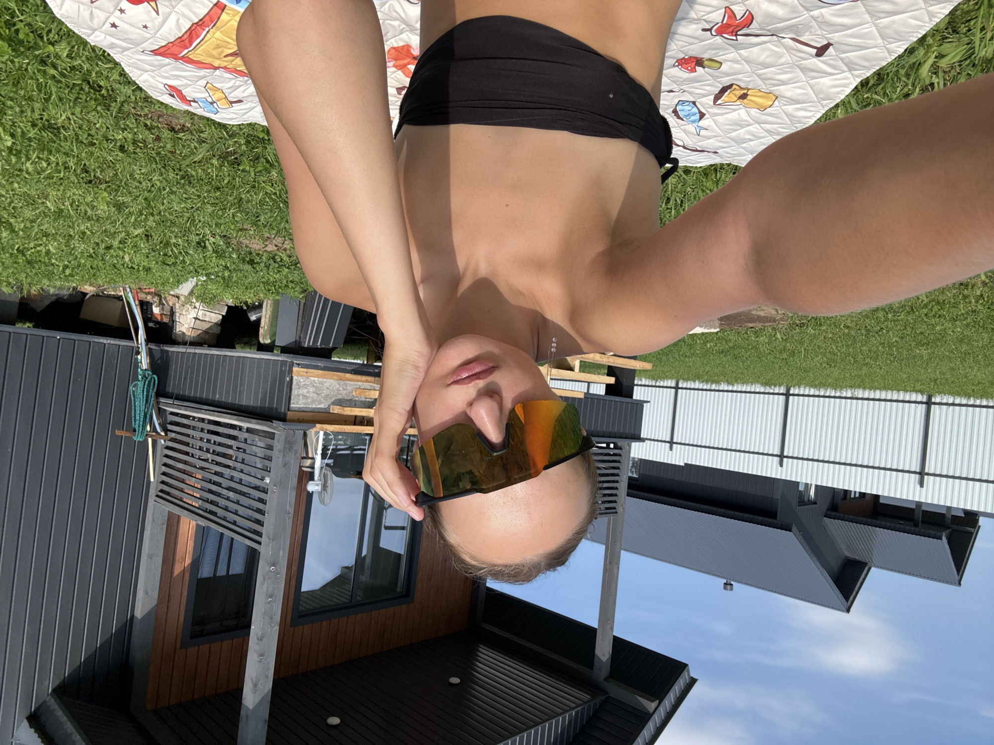 The image shows a young woman taking a selfie outdoors, sitting on a patterned blanket on a grassy area. She is wearing a black strapless bikini top and large, mirrored sports sunglasses. Her left arm is raised, holding the phone to take the picture, and her right hand is touching her face. Behind her, a modern house with dark siding and a light-colored fence is visible. The setting appears to be a residential area. The weather is sunny with a clear blue sky. The location is identified as Podshivalovo, Russia.
