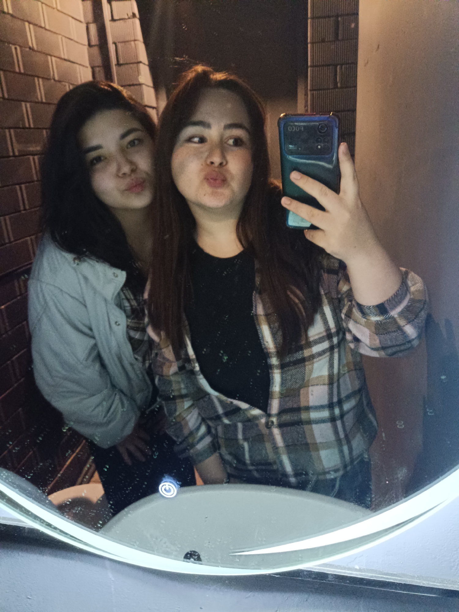 Two women are captured taking a mirror selfie indoors. The woman on the left, with dark hair, is reflected wearing a light-colored jacket over a dark top and makes a "pouty" face. The woman on the right, also with dark hair, is wearing a plaid shirt with light blue, brown, black, and white checks over a dark top. She holds a smartphone with a blue protective case to take the picture, and the inverted text "POCO" is visible on the phone. She is also making a "pouty" face, looking towards the phone.
The setting appears to be an indoor space, possibly a bathroom or a public restroom, given the prominent mirror and a partially visible white sink or countertop at the bottom. A glowing white power symbol button, likely for a mirror light or other fixture, is reflected near the bottom right of the mirror. The background reflected behind the women includes a dark, brick-patterned wall on the left and a lighter, plain wall on the right. The lighting suggests an artificial indoor environment, possibly at night or in a dimly lit area. The women's expressions convey a playful and casual mood.