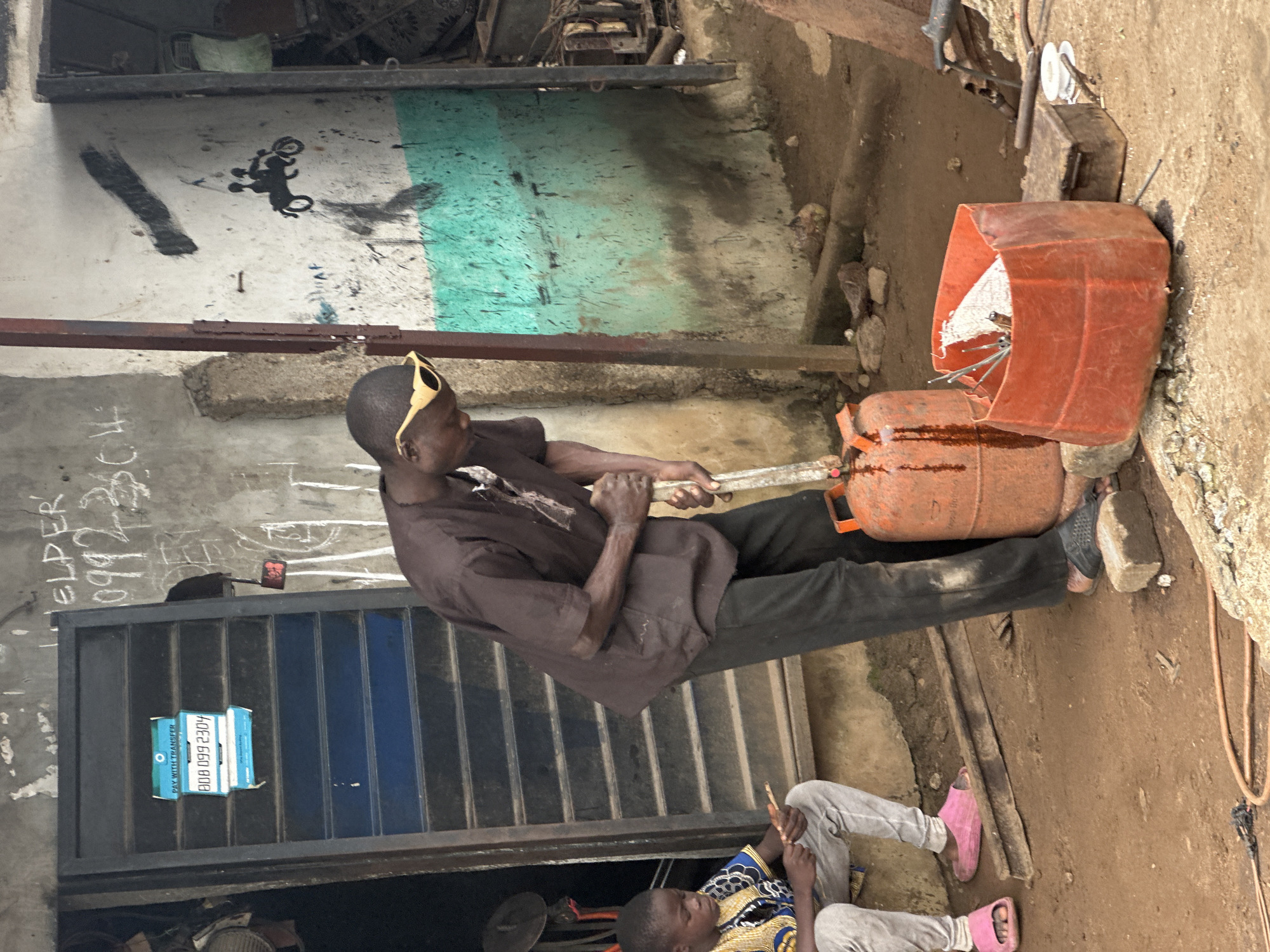 In Badagry, Nigeria, a man wearing sunglasses and a brown shirt is working outside what appears to be a workshop. He is holding a metal rod, possibly for welding, and is engaged in a task involving an orange propane tank. Next to him, an orange container holds various metal objects and tools. A young boy, dressed in a colorful shirt and light gray pants, sits nearby, holding a thin stick. The ground is dusty, and the environment suggests an outdoor, industrial setting. The workshop wall features graffiti, including what looks like a drawing of two people on a motorcycle and some text. A metal door with blue slats is visible on the left, with a sign displaying the phone number "0808 099 2304."