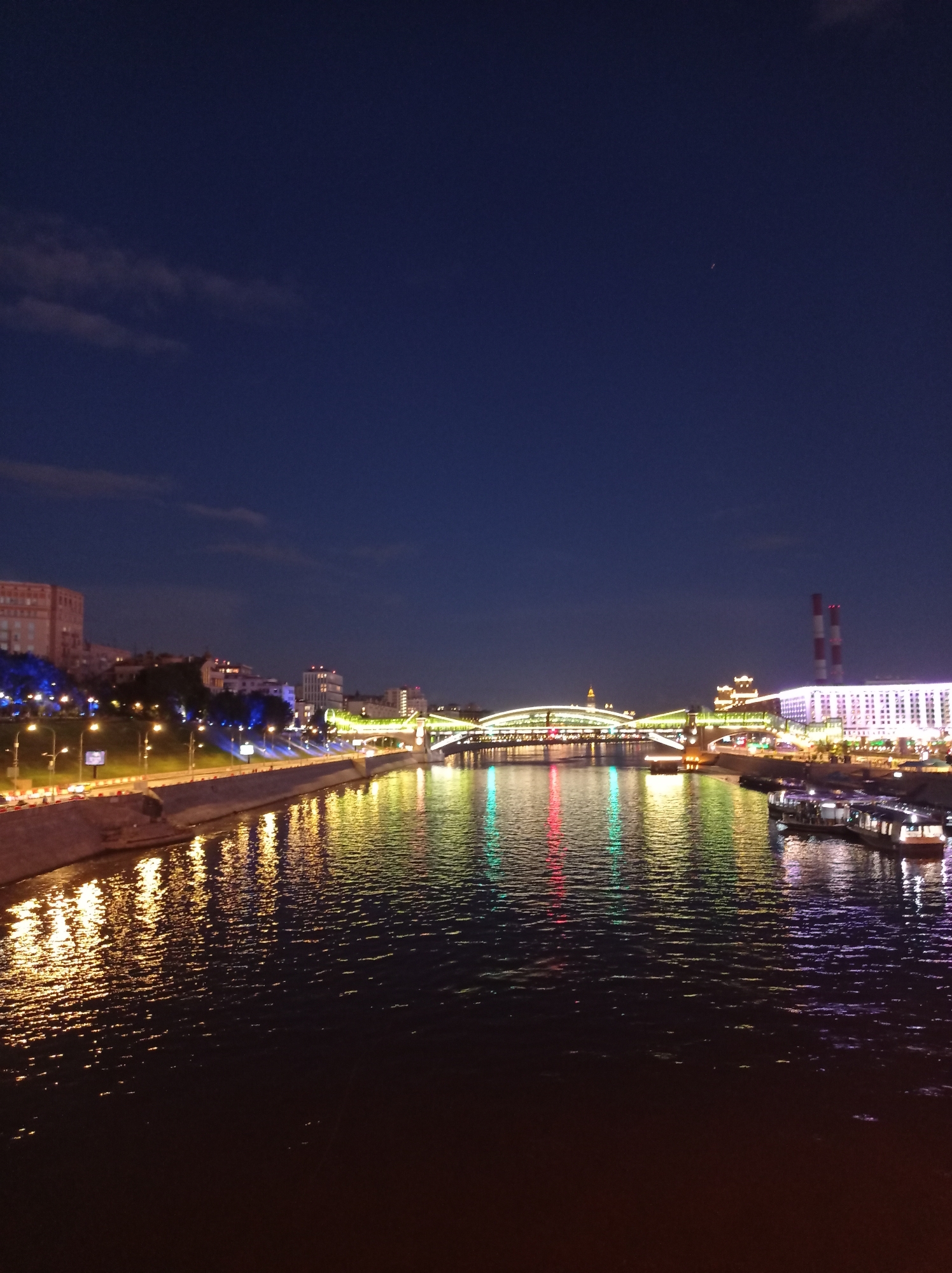 The image captures a nighttime view of the Moskva River in Moscow, Russia. The water reflects the colorful lights of buildings and bridges along the embankment. On the left bank, buildings are illuminated with warm, yellow lights, and trees are lit with blue lights. A street with traffic lights and cars is visible along the embankment. In the center, a long bridge with green and yellow lights spans the river. On the right bank, a large white building is brightly lit with purple and pink lights, and two red and white smokestacks rise above it. Several boats are docked along the embankment. The sky is a deep blue with a few faint clouds and a tiny light, possibly an airplane or star, visible in the upper right portion of the frame. The overall scene depicts a vibrant urban landscape at night, with the river acting as a mirror for the city's illuminated architecture.