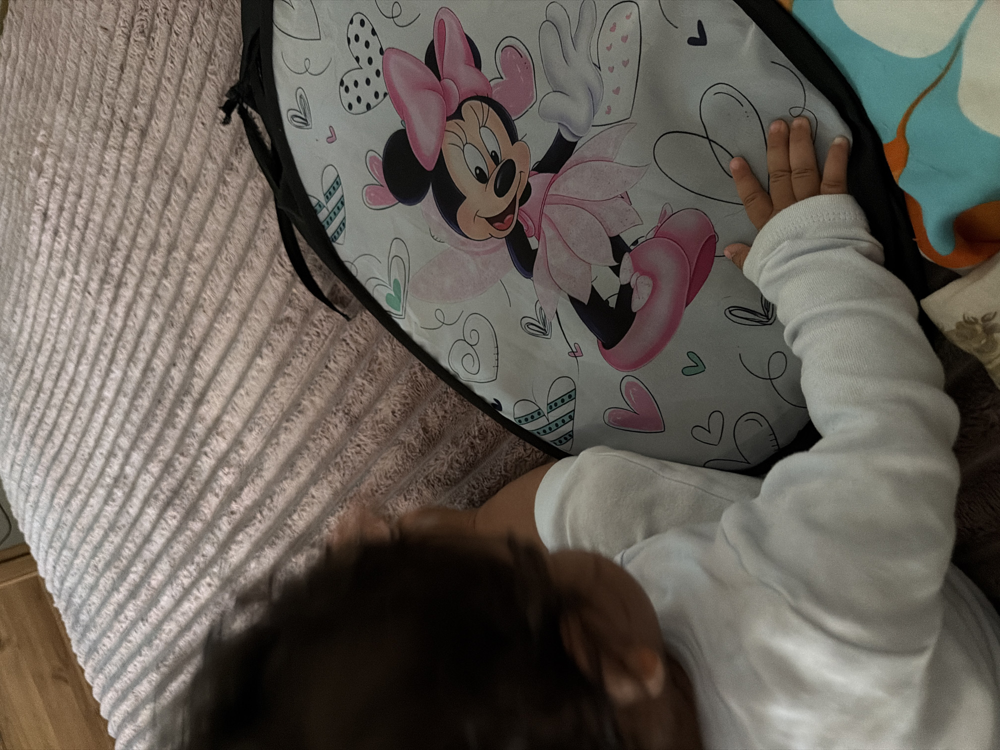 A young child, with dark hair and wearing a white long-sleeved shirt, is seen from the back and side, looking down at a mat featuring Minnie Mouse in a pink outfit, surrounded by hearts. The child's left hand rests on the mat. The background consists of a textured, pinkish-grey surface, possibly a bedspread, and a wooden surface on the far left. The scene appears to be indoors, in a bedroom, in Razhevo Konare, Bulgaria.