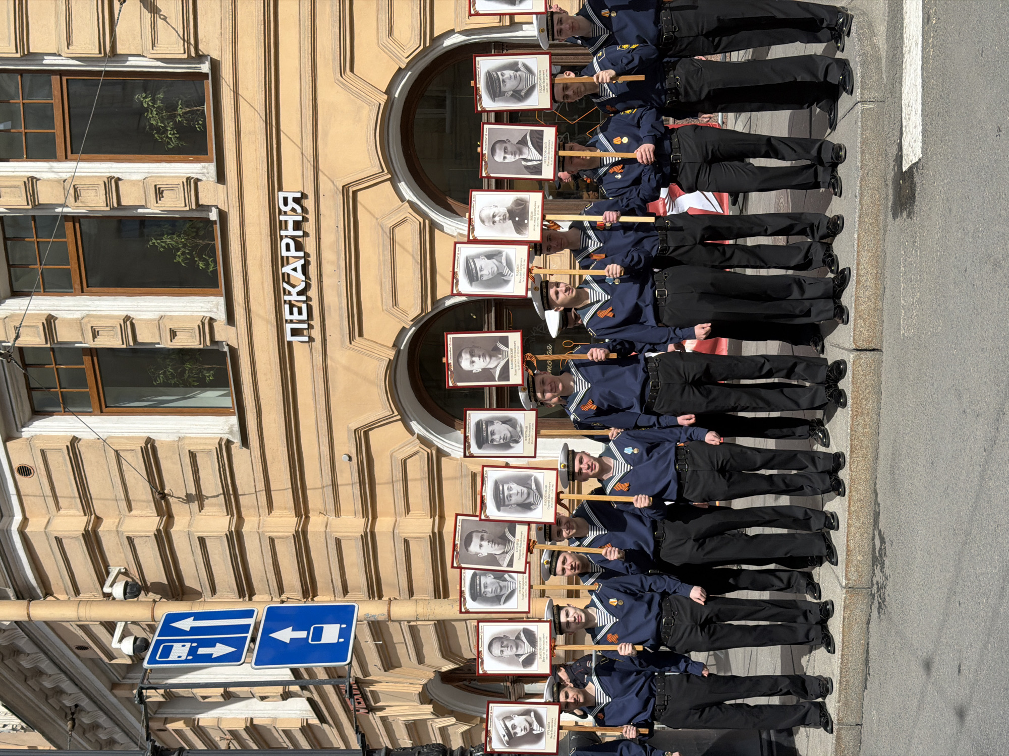 The image shows a group of young men dressed in navy uniforms standing in a row, each holding a portrait on a stick. The portraits appear to be of historical figures, possibly related to military service. The young men are standing on a street next to a building with a sign that reads "ПЕКАРНЯ" (Bakery) in Russian. Above the street, there are road signs indicating directions for vehicles. The architectural style of the building suggests it is located in Saint Petersburg, Russia.