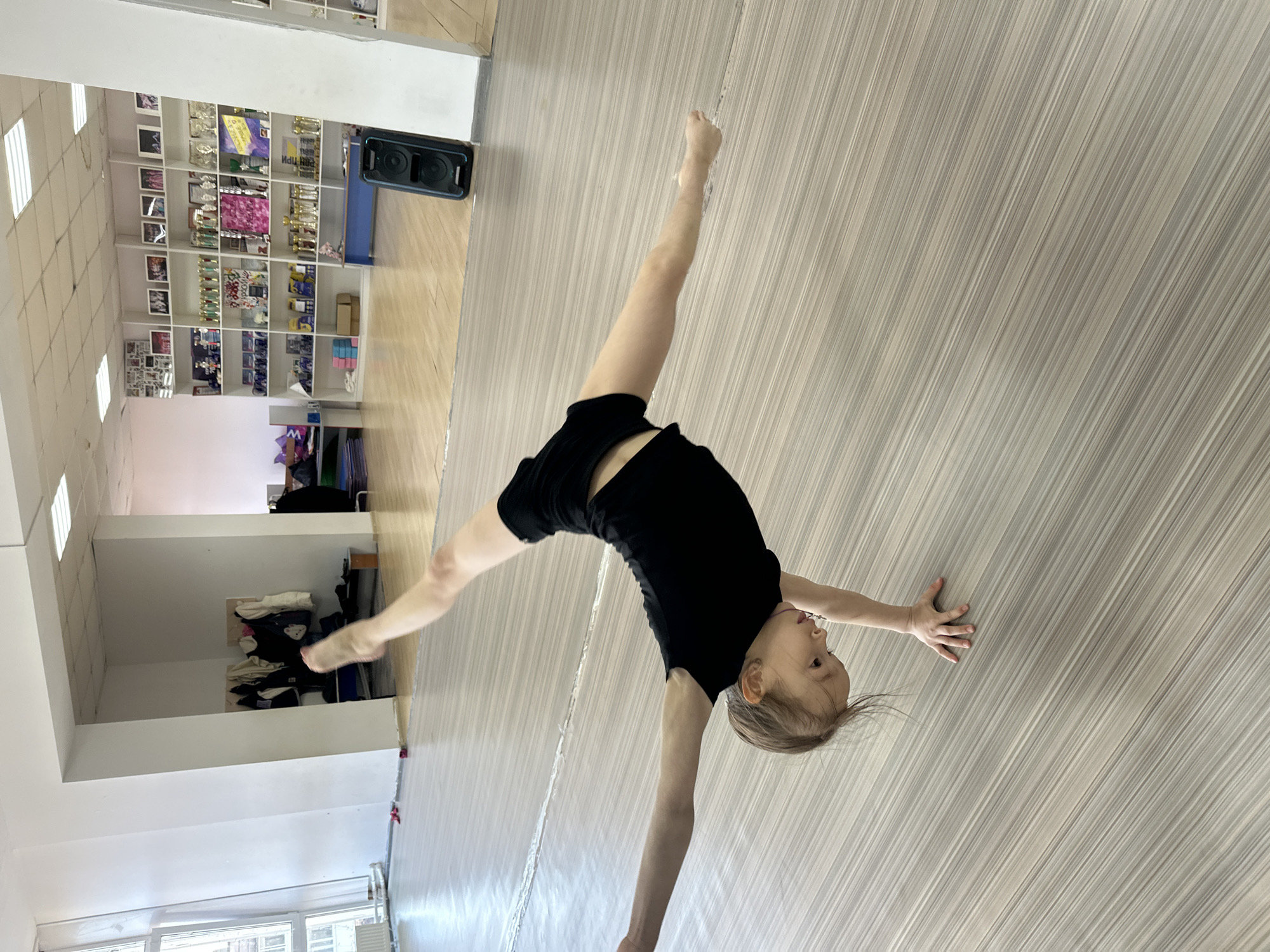 A young girl in a black leotard is performing a handstand split on a light gray floor with linear patterns. The location appears to be an indoor space, possibly a dance studio or gymnasium, in Votkinsk, Russia. In the background, there is a shelf filled with trophies and framed pictures. A black speaker sits on a wooden ledge above a blue bench. A small window can be seen in the bottom left corner of the image.