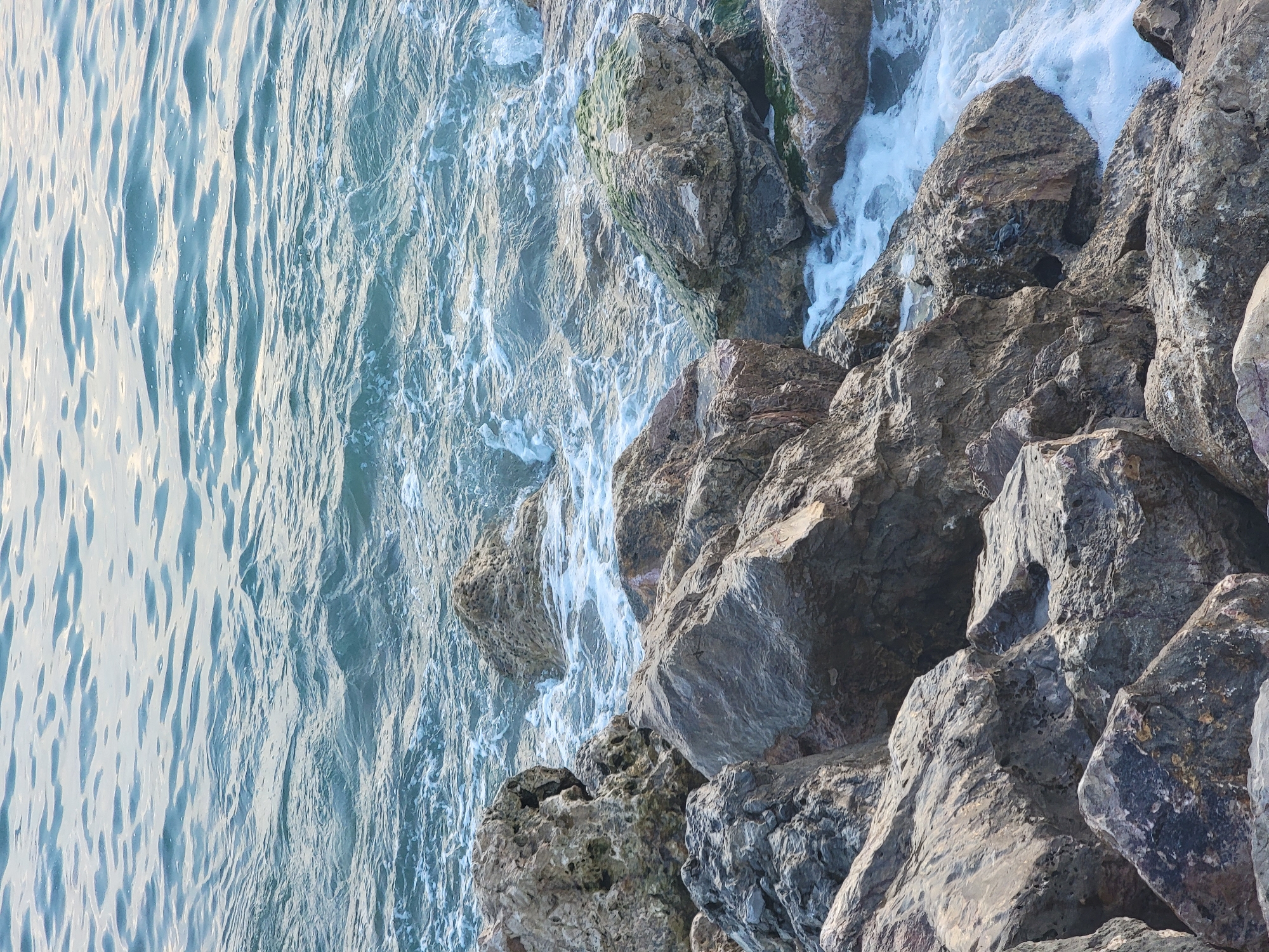 This is a photograph of the sea crashing against a rocky shoreline in Quarteira, Portugal. The water is a deep blue with white foam as it hits the rocks. The rocks are varied in size and color, with some covered in green moss. The scene appears to be a natural and undisturbed environment.