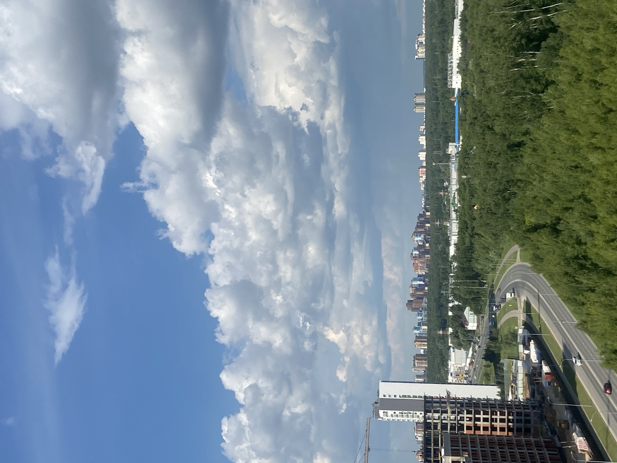 An aerial, high-angle, full outdoor shot captures a cityscape of Kazan, Russia, under a partly cloudy sky. A large, partially constructed building with exposed rebar and concrete dominates the left side of the frame, accompanied by a construction crane. Adjacent to it stands a finished, multi-story building with a white facade. A highway with several vehicles, including cars and trucks, curves through the scene, bordered by a dense forest of green trees. In the distance, a cluster of modern residential buildings forms the city skyline, nestled amongst more greenery. The sky is a vibrant blue with scattered white and grey clouds, some of which appear dark and heavy.