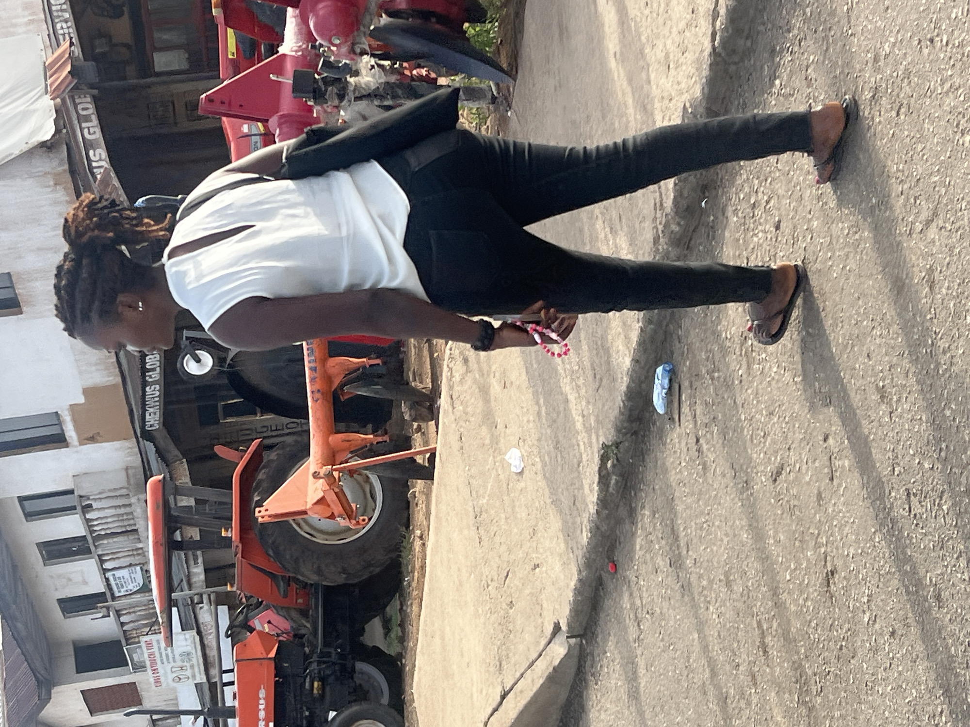 In Gura Top, Nigeria, a woman walks on a concrete road beside a commercial building identified as "Cheikhus Global Services." She wears a white shirt, black jeans, and flip-flops. She carries a black purse. An orange tractor is parked nearby, partially obstructing the building. Scattered litter is visible on the road.