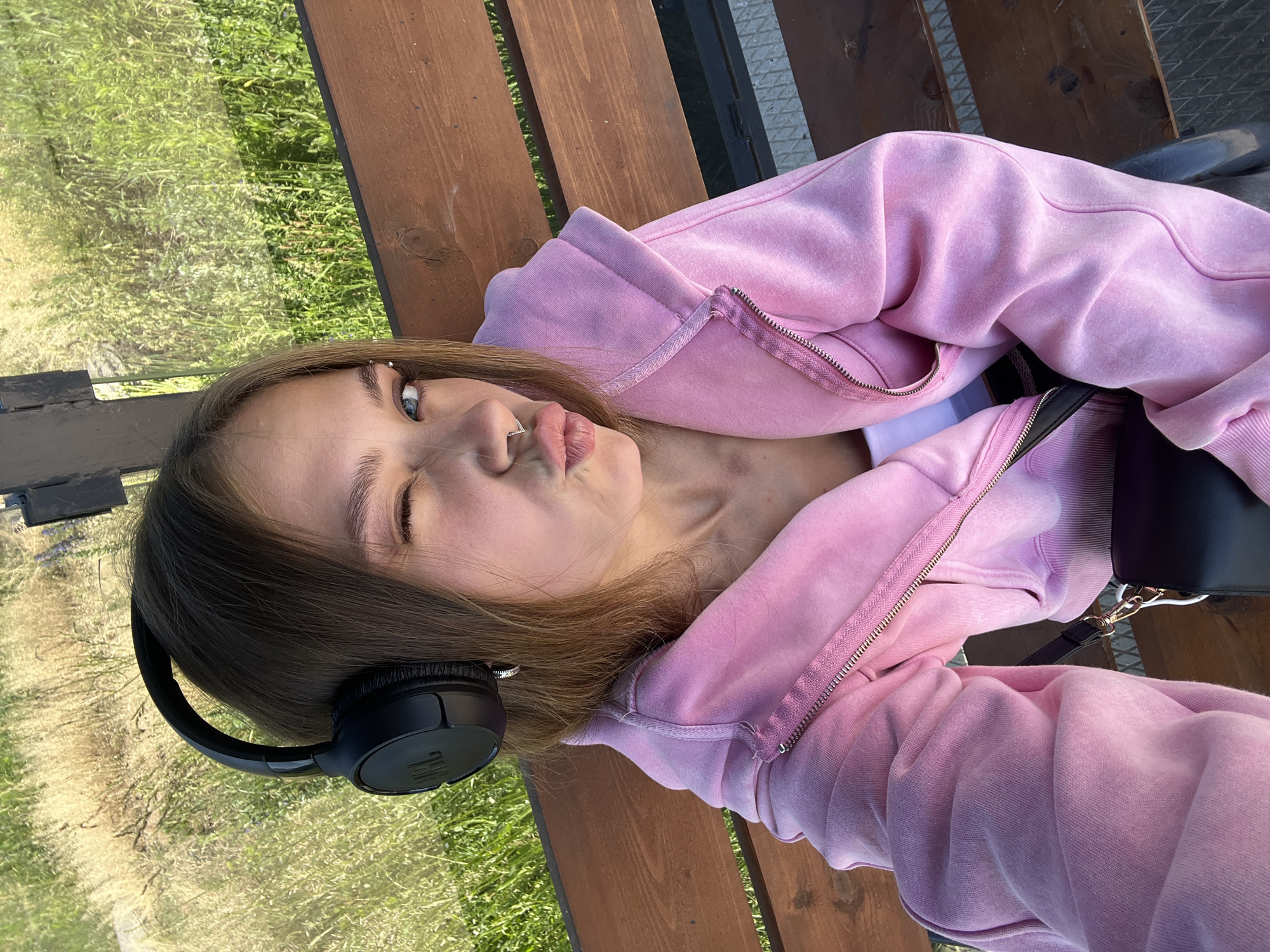 A young female wearing a pink hooded sweatshirt and black headphones is sitting on a wooden bench. She is looking at the camera and winking. Her lips are pursed, and she has a nose ring. The background consists of green grass and some dried vegetation. This scene is located in Chaikovsky, Russia.