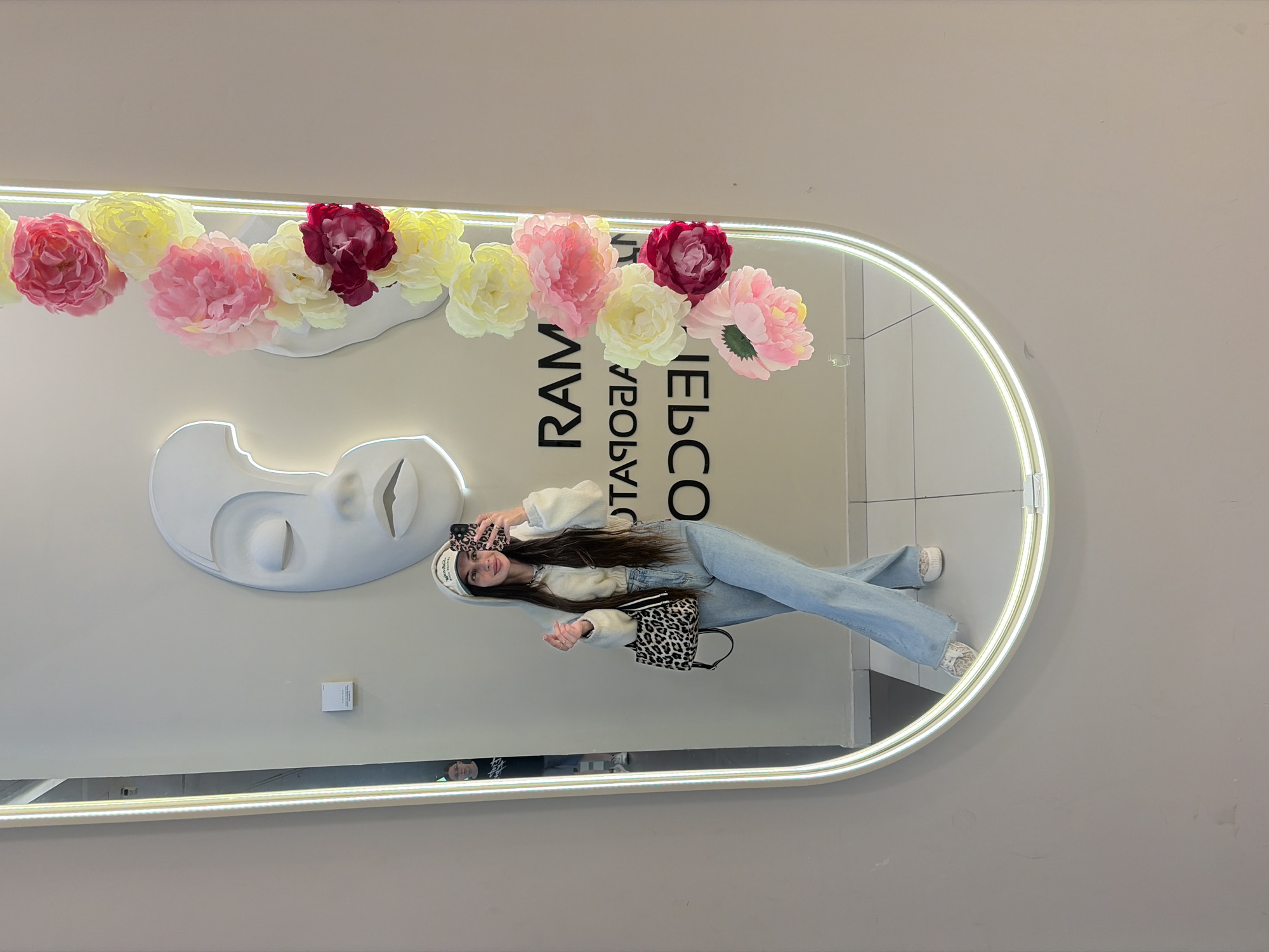 A young woman with long dark hair, wearing a white baseball cap, a cream-colored cropped hoodie, and light blue flared jeans, takes a mirror selfie. She is holding a cell phone with a leopard-print case in her right hand and a leopard-print handbag in her left. She is standing in front of an oval mirror with an illuminated rim. The mirror reflects the woman, a person in the background wearing a dark shirt and glasses, and part of the environment. The wall behind the mirror features a large white abstract sculpture of a face and a string of artificial flowers in shades of pink, yellow, and burgundy cascading down the right side of the mirror. Black lettering on the wall reads "RAM" and "HEBCC", which appears to be reversed lettering for "RAMP" and "CHECK" or similar. The scene appears to be an indoor setting, possibly a retail store or a fashion boutique, based on the decor and the reflected signage. No specific landmarks or street views are visible to definitively place it within Saint Petersburg, Russia, however, the signage could indicate a business located there.