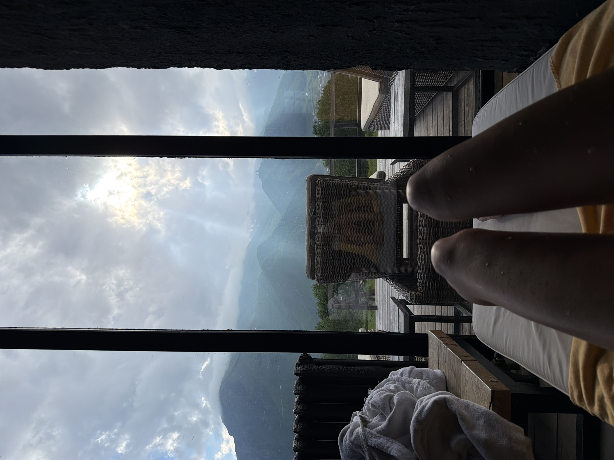 The image shows a person's legs resting on a lounge chair, with a view of mountains and a cloudy sky through a window. The scene is set outdoors, likely on a balcony or patio, with outdoor furniture visible. A white towel is draped over the side of the lounge chair. The lighting suggests it might be daytime, possibly early morning or late afternoon, given the soft, diffused light. The weather appears overcast with patches of sunlight breaking through the clouds. The mountainous landscape suggests a location in a rural or remote area, consistent with the provided location context of Stepantsminda, Georgia. The overall mood conveyed is one of relaxation and enjoyment of a scenic view. There is no visible text in the image.
