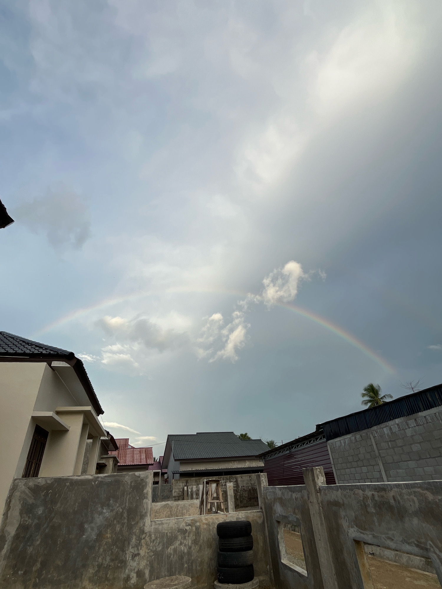 The image depicts a residential area in Lampeuneurut Gampong, Indonesia. A double rainbow arches across a cloudy sky, with the brighter arc visible through thinner clouds and the fainter arc to its right. In the foreground, a partially constructed concrete wall encloses a yard. Beyond the wall, several houses with tiled roofs are visible, including a modern house with a beige facade on the left and a house with a dark gray roof in the center. The background shows a line of corrugated metal roofing and a brick wall. A palm tree is visible to the right of the frame. The scene appears to be after a rain shower, as suggested by the presence of the rainbow. No people are visible in the image.