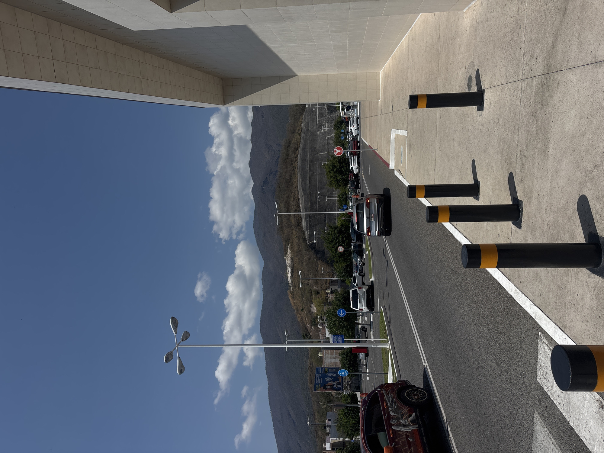 The image shows a street scene in Chilpancingo, Mexico. A line of parked cars is visible on the right side of the road, next to a curb lined with black and yellow bollards. A dark-colored car is partially visible in the foreground on the right. In the background, mountains rise into a blue sky with white clouds. A street light with a four-headed fixture is visible on the left side of the road. A large, light-colored building is partially visible on the top left corner of the image. Various traffic signs are visible along the street.