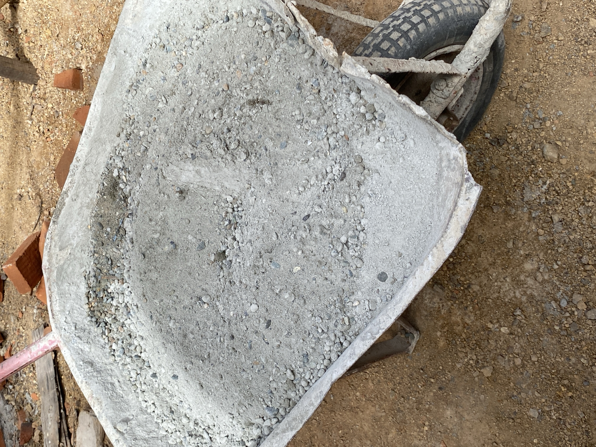 A close-up, high-angle shot captures a weathered, galvanized metal wheelbarrow filled with a mixture of dry cement and small stones. The wheelbarrow's rim is chipped and worn, with visible signs of past use. The dry mix forms a shallow mound within the basin, with larger stones concentrated in a few areas. The wheelbarrow is positioned on a ground surface composed of dirt and small pebbles, indicative of a construction or outdoor work environment. In the background, scattered red bricks and pieces of wood can be seen, further suggesting a construction site. The lighting suggests it is daytime, with some shadows indicating the sun's position. The scene appears to be located outdoors.