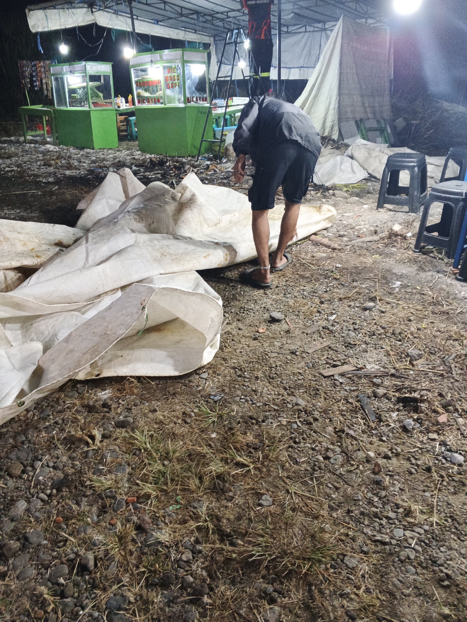 The image depicts an outdoor scene at night, likely a market or food stall area in Sukabumi, Indonesia. In the foreground, a person wearing black shorts and a grey jacket is bending over a pile of large white tarps or canvas sheets. The ground is covered with gravel, dry grass, and debris.
In the background, two bright green food stalls are visible, illuminated by overhead lights. One stall has signage that appears to say "MIE AYAM" (chicken noodles). There are also plastic chairs, some black and some blue, scattered around the area. A ladder leans against a structure behind the stalls, and a tent-like structure made of white fabric is also present. The overall atmosphere is dimly lit and appears to be in a somewhat unkempt outdoor environment.