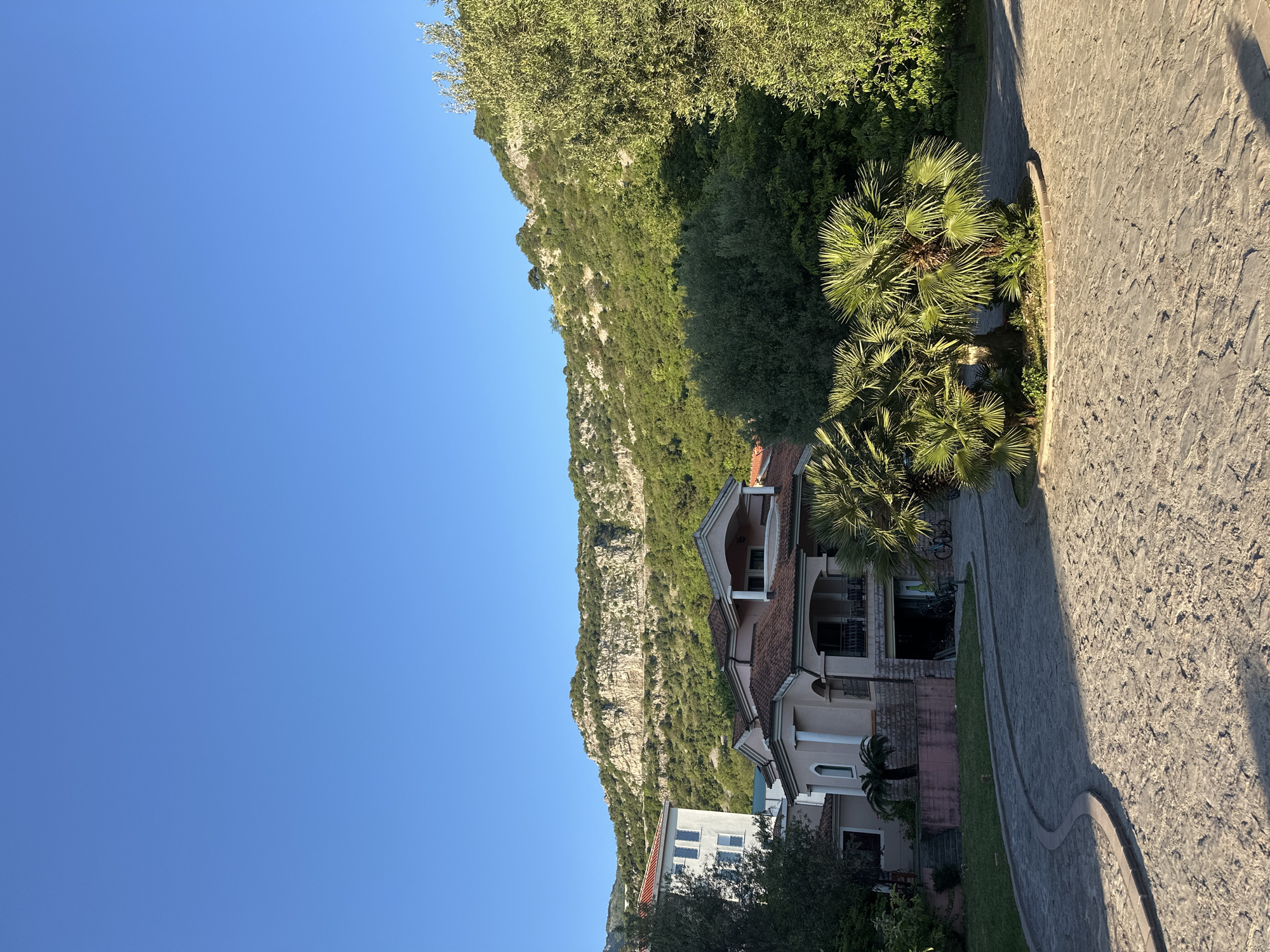 The image shows a residential area in Budva, Montenegro. In the foreground is a stone-paved road, with trees and vegetation on the right. A light-colored multi-story building with a red-tiled roof is visible, with balconies and arched openings. A palm tree stands next to the house. A mountain covered with green trees and vegetation rises in the background, with a clear blue sky above. Another white building with a red-tiled roof is visible at the bottom left of the image. The lighting suggests a sunny day.
