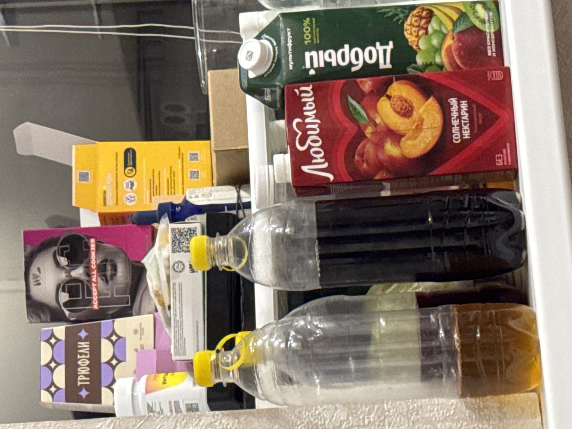 The image shows a shelf with various items, including two large plastic bottles filled with dark liquid, a carton of "Любимый" peach nectar, a carton of "Добрый" multifruit juice, and several boxes and pill bottles. The items are arranged in a somewhat cluttered manner. The background appears to be a wall with some shelving or cabinets, and the overall lighting suggests an indoor setting. The image does not provide any specific visual cues that would allow for determination of its location within the city of Saratov, Russia. There are no people or actions depicted. The focus is on the still life arrangement of products.