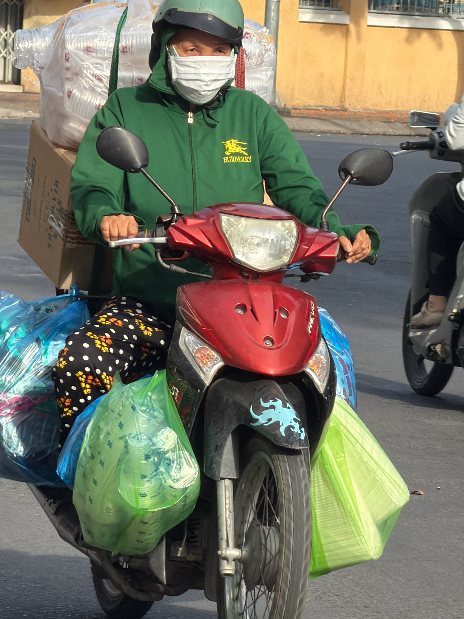 A person wearing a green jacket, a green helmet, and a white face mask rides a red scooter in Nha Trang, Vietnam. The scooter is laden with several large, clear plastic bags filled with items and a cardboard box is strapped to the back of the scooter. The person's legs are visible, wearing black pants with a white polka dot and floral pattern. The background shows a building with a yellow facade. The scooter appears to be in motion, as indicated by the blurred motion of the wheels and the environment.