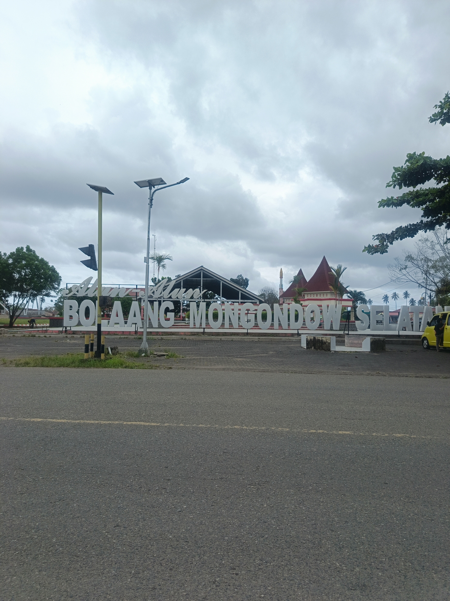 The image captures a wide-angle view of the welcome sign for "BOLAANG MONGONDOW SELATAN" in the city of Tabilaa, Indonesia. The sign, made of large white letters, is prominently displayed in the foreground, with the words "Alun Alun" appearing above it in cursive. Behind the sign, a paved area leads to a building with a distinctive red roof and a minaret, suggesting it might be a mosque or a community center. Several palm trees dot the landscape. Two solar-powered streetlights stand near the sign, one of which is positioned in front of it. On the right side of the image, a yellow car is parked, with a person partially visible inside. The environment appears to be an open, public space, possibly a town square or entrance to a district. The sky is overcast with heavy clouds. The road in front of the sign shows a yellow dividing line.