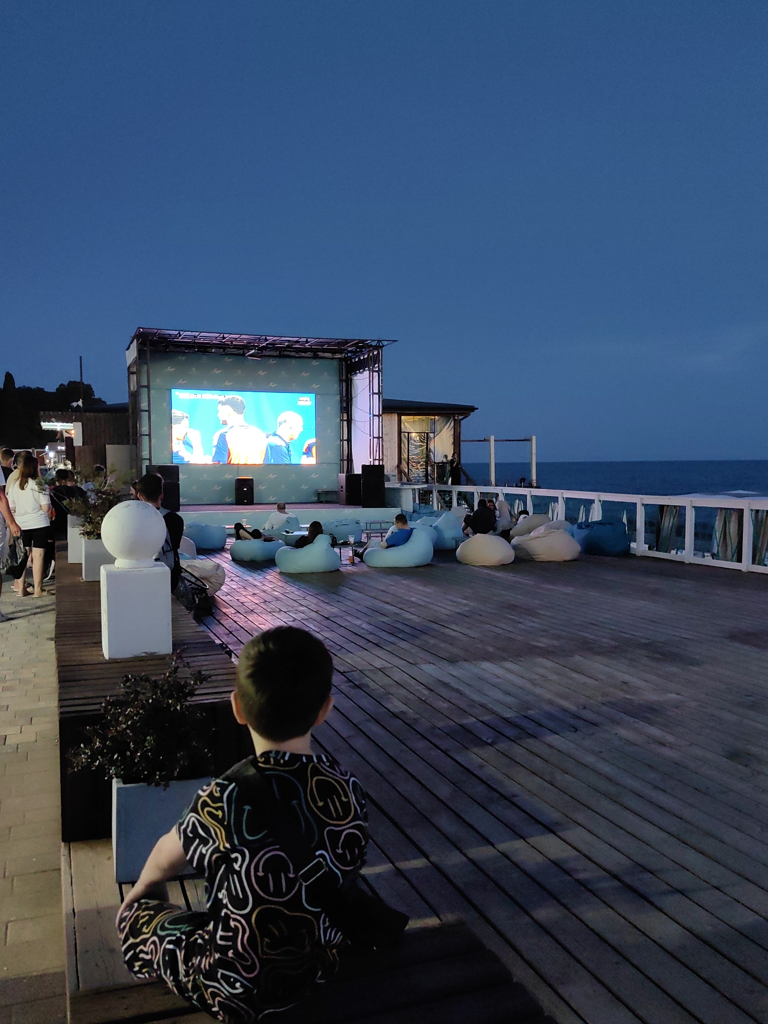The image depicts an outdoor cinema setting at dusk, likely on a pier or boardwalk overlooking the Black Sea. The location appears to be the resort city of Sochi, Russia, given its coastal setting and typical entertainment infrastructure. A large screen is set up on a stage, displaying what appears to be a sporting event or broadcast. In front of the screen, a crowd is seated on beanbag chairs, facing the display. To the left, a group of people are standing, some looking towards the screen and others seemingly engaged in conversation. A young boy is in the foreground, facing away from the camera, seated on a bench. The overall atmosphere is relaxed, with a sense of community gathering to watch the event. The lighting suggests it's evening, with the sky a deep blue and artificial lights illuminating the scene. The presence of the sea and a railing with curtains suggests a seaside promenade or entertainment area common in resort towns.