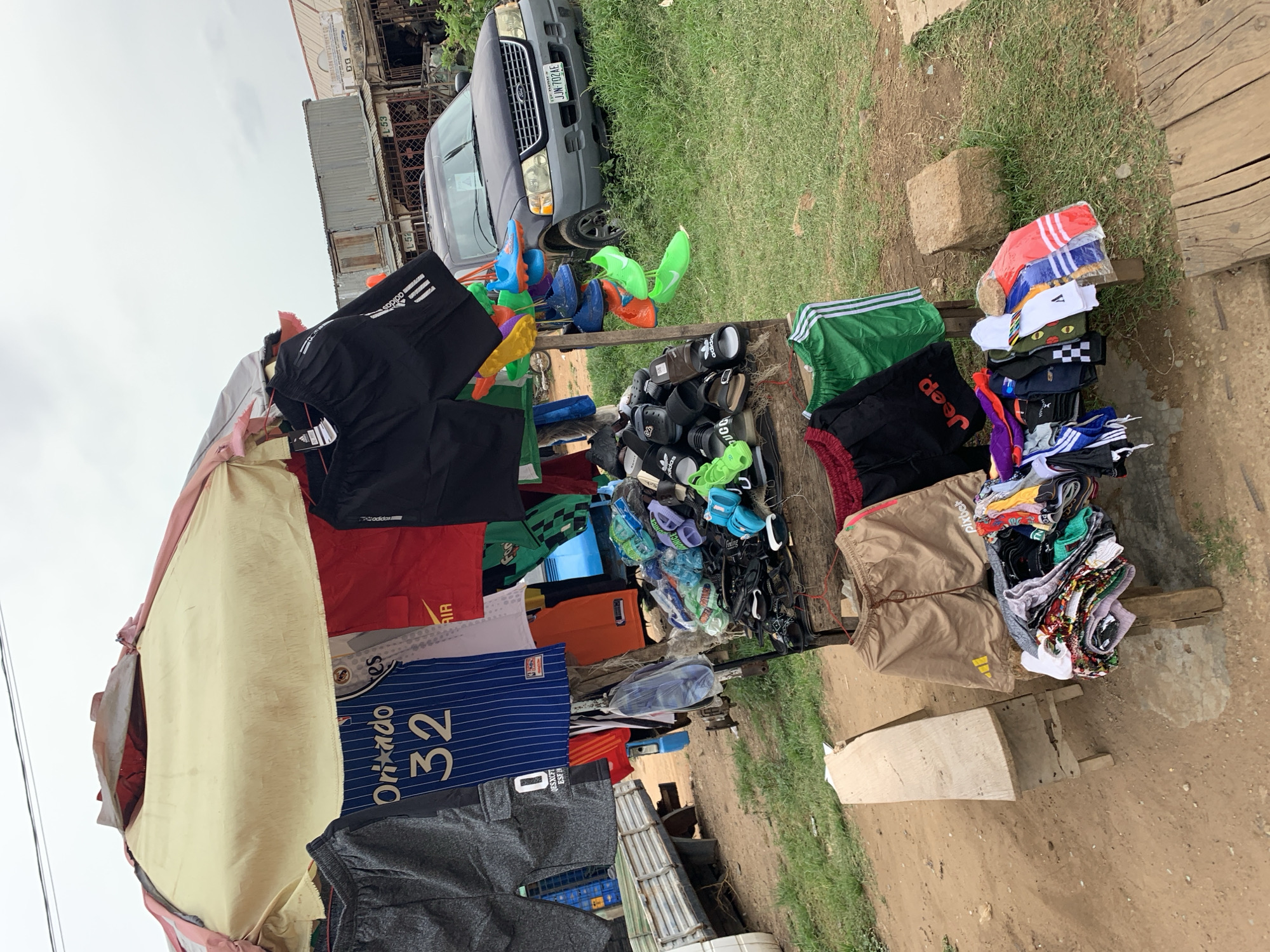 The image captures an outdoor market stall in Jos, Nigeria, displaying a variety of clothing and footwear. Merchandise is arranged on makeshift wooden tables and hanging racks under a beige fabric canopy.
The primary subjects are numerous items of clothing and shoes. There are various sports shorts, including black Adidas shorts with three white stripes, green striped shorts, red shorts with "AIR" text, tan shorts featuring "pixbet," and black shorts with "Jeep" lettering. Several soccer jerseys are visible, notably a blue and white striped jersey with "Orlando 32." A prominent collection of footwear, mainly sandals and slippers, is stacked on tables, some clearly branded "Adidas" or with text like "NICE" and "DONICE." Colorful socks, some bearing an "Adidas" logo or a "V" emblem, are also neatly piled.
In the background, a dark grey Ford Explorer SUV is parked on a grassy patch, bearing the Nigerian license plate "JJN-702AE." Behind the vehicle, several corrugated metal containers, possibly market stalls or storage units, are visible, marked "L-53" and "L-54." Distant buildings show partial text such as "MASON MUIST WENT DO." The ground beneath the stall and car is a mix of dirt and grass. The sky is overcast, indicating daytime.
The scene depicts a typical roadside vending operation, implying commercial activity where goods are presented for sale. While no people are directly visible, the organized display suggests an active marketplace.