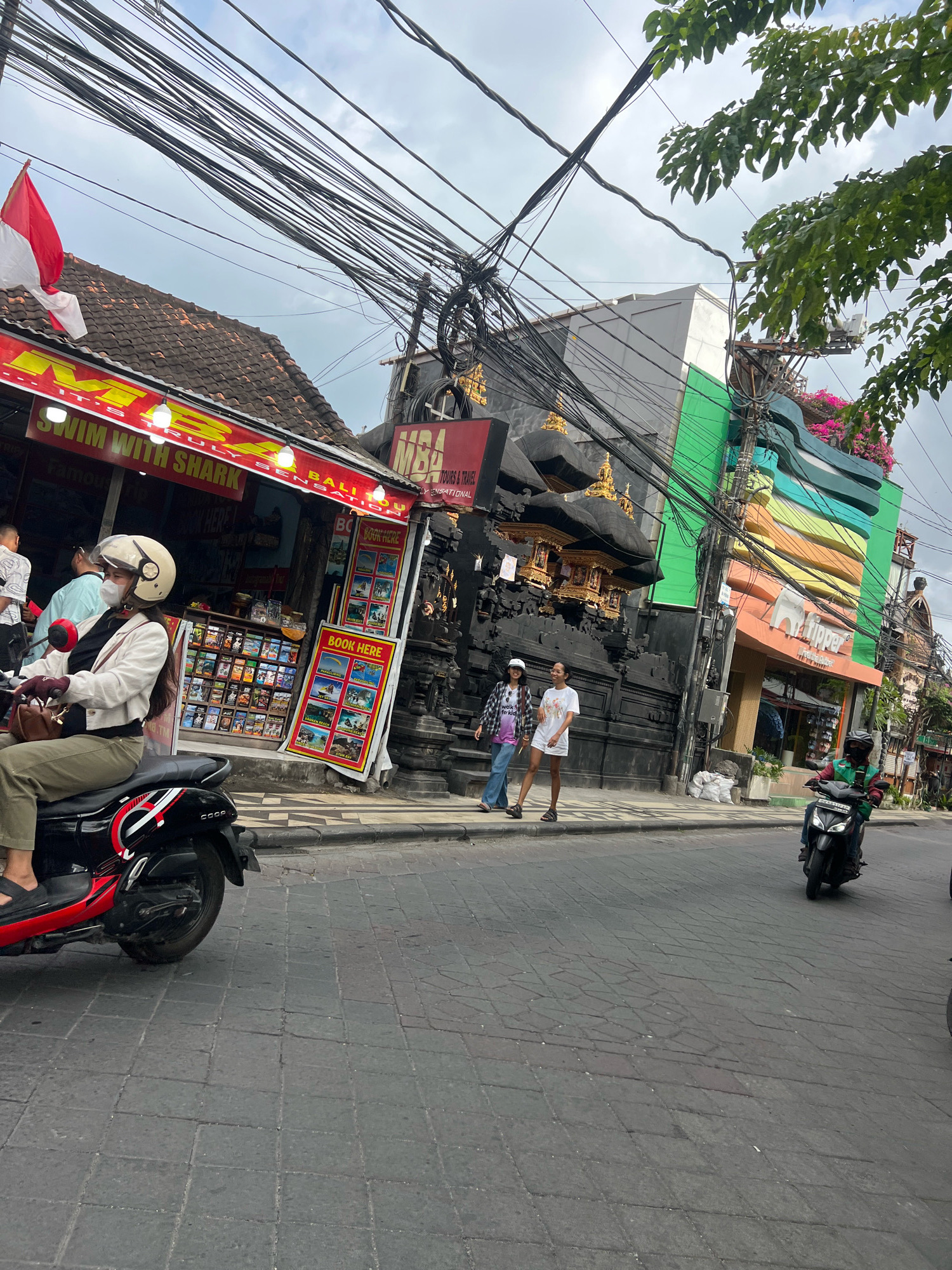 In Kuta, Indonesia, on a cloudy day, a street scene unfolds with a focus on a busy sidewalk and roadway. A woman wearing a white helmet, jacket, and olive green pants rides a black and red scooter, her left hand on the handlebar and her right hand gloved and resting on her leg. She is looking ahead, her face obscured by a face mask and helmet visor. Behind her, a red and white Indonesian flag is visible.
The sidewalk is lined with shops. One shop, with a red awning, advertises "SWIM WITH SHARK" and "BOOK HERE," displaying various tour brochures. Another building features a dark, ornate facade with gold accents, and a brightly colored facade with the word "lipper" visible.
Two women stand on the sidewalk, one in a plaid shirt and jeans, the other in a white t-shirt and shorts. Further down the street, a person on a scooter in a green shirt rides past. The roadway is paved with dark cobblestones, and the sky is filled with a dense network of overhead electrical wires.