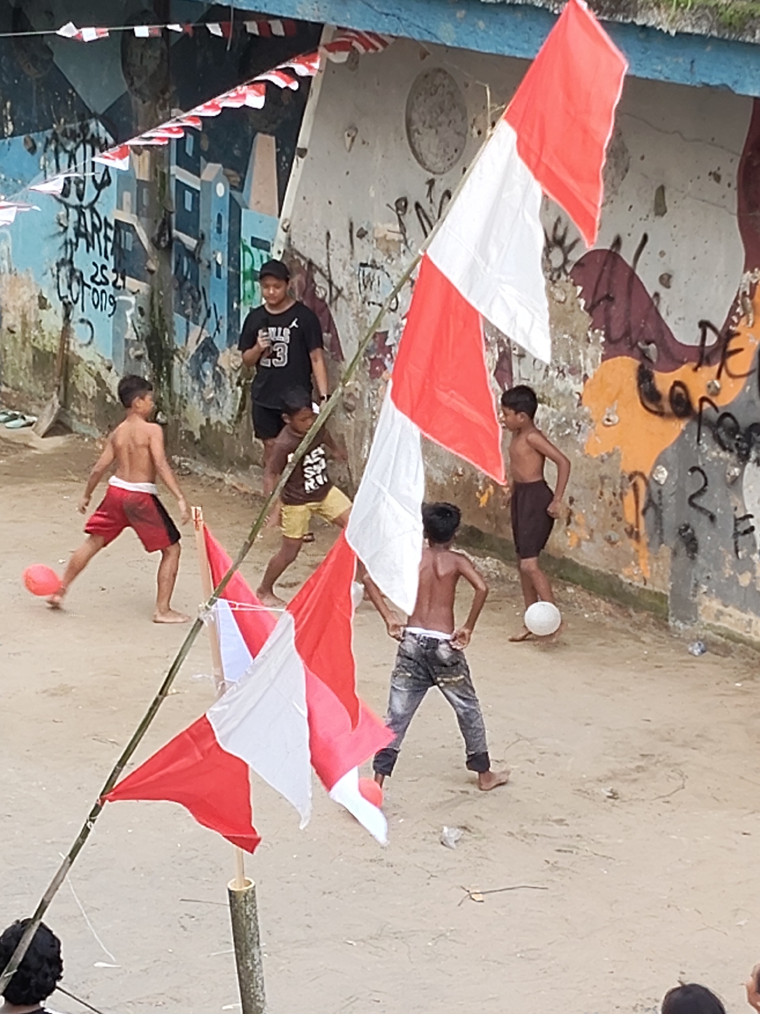 A high-angle, medium shot captures several young boys playing with soccer balls in a dusty, outdoor area, possibly a street or an open lot, near a graffiti-covered wall in the City of Medan, Indonesia. The wall features vibrant, though somewhat faded, murals and tags, with a blue and white striped awning overhead decorated with small red and white flags. The ground is a mixture of dirt and sand, with scattered debris.
In the foreground, two boys are actively engaged in play. One, wearing dark jeans and a black t-shirt with the number "23" visible, stands to the left, appearing to be in mid-action. Another boy, shirtless and wearing red shorts with black trim, is to the left of center, his right foot poised to kick a small, reddish-orange soccer ball.
Further back, another boy, shirtless and in dark shorts, is near the wall, his left foot about to kick a white soccer ball. Behind him, another boy, shirtless and wearing brown shorts, looks on. A fifth boy, wearing a dark t-shirt with "BULLS 23" and black shorts, stands further back, observing the game. Two Indonesian flags, one red and white, and another white, are prominently displayed on bamboo poles in the foreground, suggesting a patriotic or celebratory event. The overall atmosphere appears to be one of casual, energetic play in a somewhat neglected urban environment.