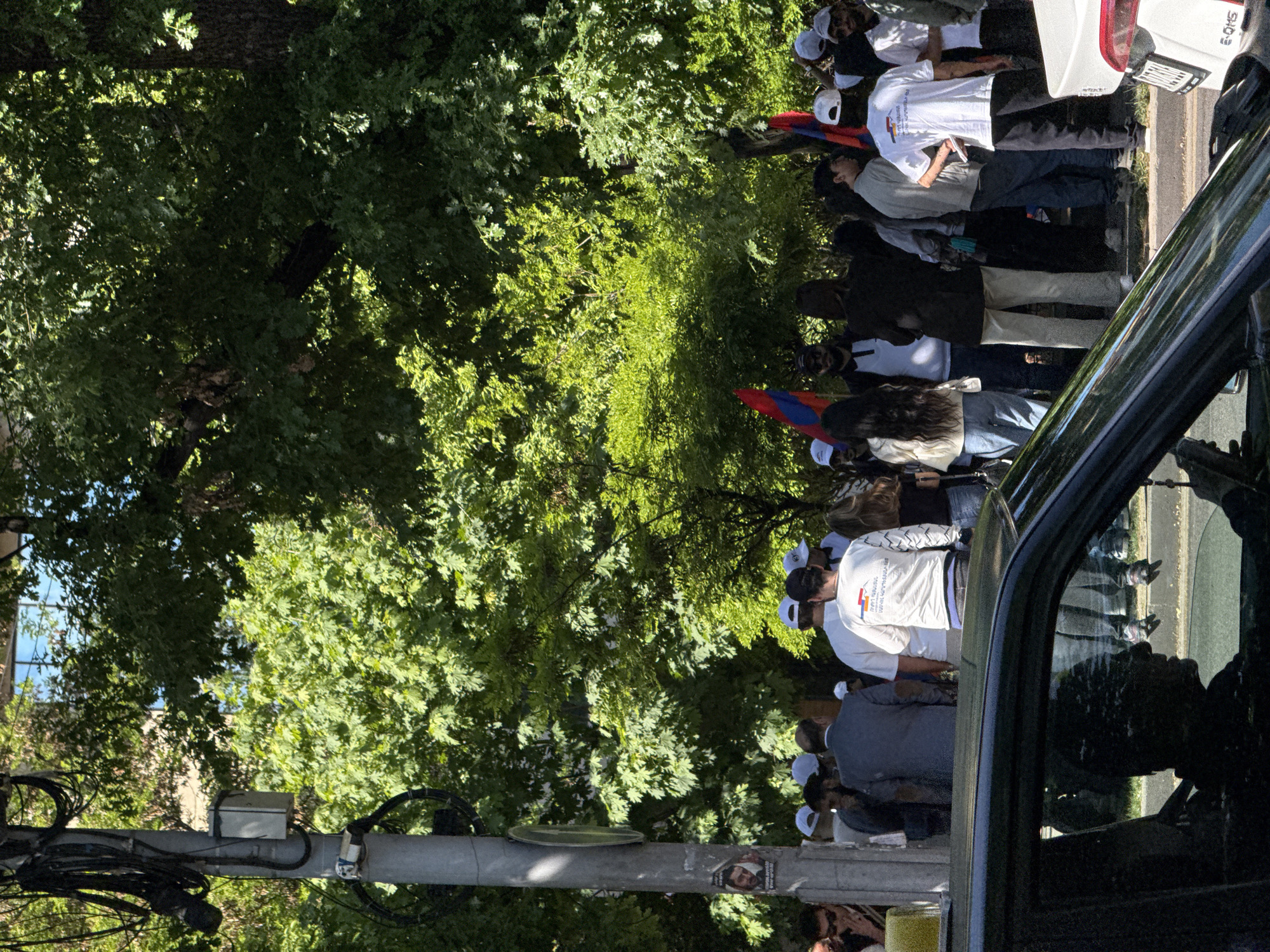 A small group of people, some wearing white shirts and baseball caps, stand on a sidewalk in Yerevan, Armenia. They are lined along a sidewalk next to a street. In the middle of the group, someone holds a flag with three horizontal stripes of red, blue, and orange. A parked white vehicle and a black vehicle are visible to the right of the group. Trees line the background of the scene. A utility pole with several wires is to the left of the image.