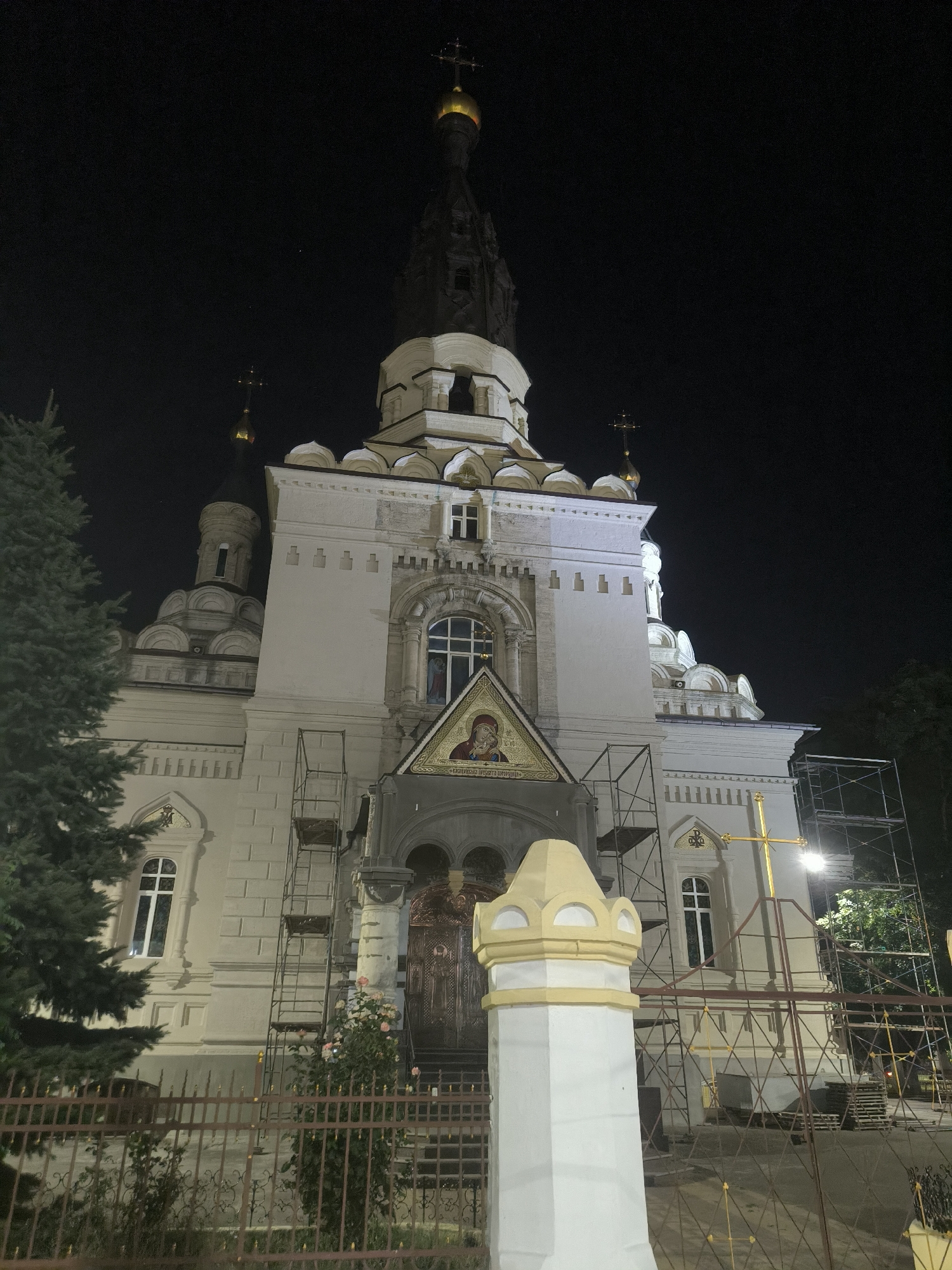 The image shows the exterior of a white, multi-domed church at night. The church has a tall, dark spire topped with a golden dome and cross. There are also several smaller domes and crosses visible. The facade of the church is white with decorative architectural elements. Scaffolding is present on the right side of the building. A large, illuminated golden cross stands in front of the scaffolding. A wrought-iron fence encloses the area in the foreground, with a decorative white pillar to the left. A large evergreen tree is visible on the left side of the image. The sky is dark, indicating nighttime. The church is identified as the Church of St. Nicholas in Mykolaiv, Ukraine. There are no people visible in the image.