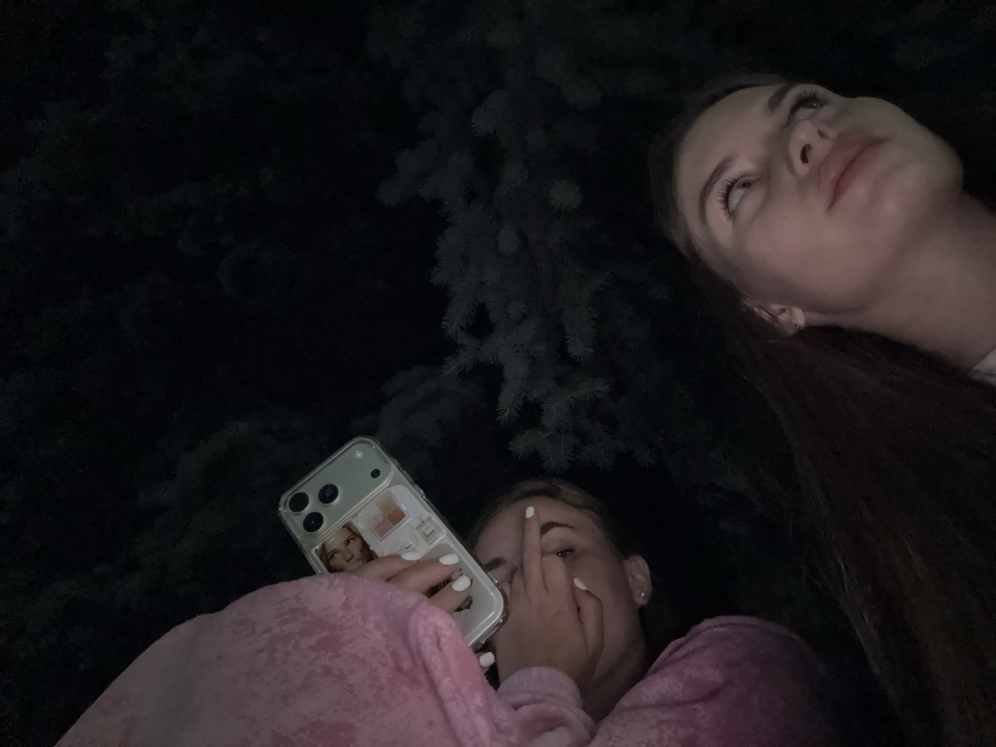 Two young women are seen at night, outdoors, with dark foliage in the background. The woman in the foreground is partially visible, wearing a pink hooded garment. She is holding a smartphone in her left hand, displaying what appears to be a photo of a person on the phone case. Her right hand is raised to her face, obscuring her mouth and nose, with her middle finger extended. Her expression is difficult to fully discern due to the hand. The woman in the background is looking upwards and to the right, with her face illuminated by an unseen light source. Her expression appears thoughtful or wistful. The image is dark, suggesting it was taken after sunset or before sunrise. No specific weather conditions are evident. The presence of foliage suggests a natural outdoor setting, possibly a park or garden. The location context is Dnipro, Ukraine. No visible text is present in the image.