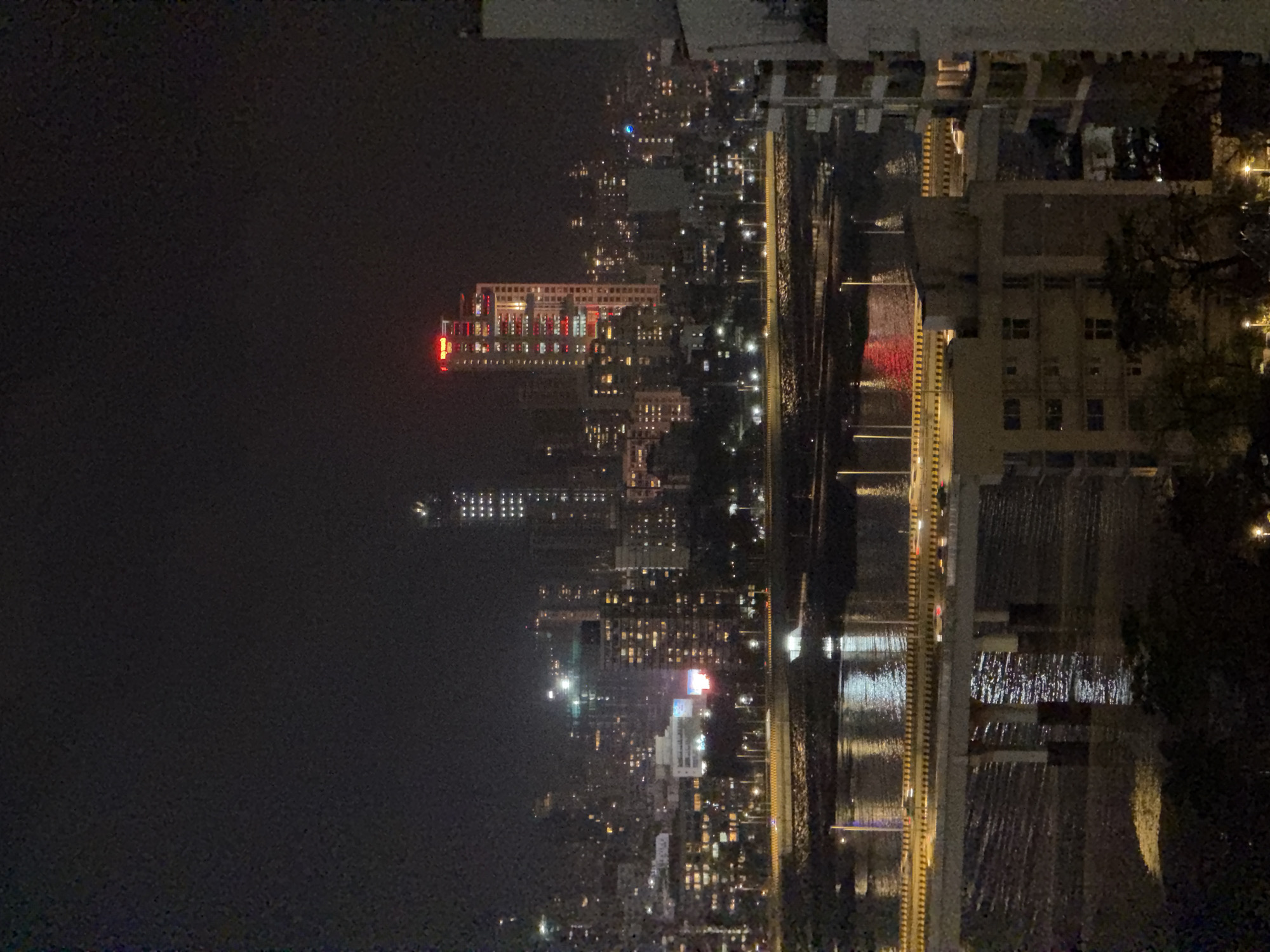 This is a nighttime aerial view of a city skyline, likely Mumbai, India, featuring a body of water in the foreground and a densely packed urban landscape beyond.
The foreground shows a wide body of dark water, reflecting the lights from the city and a bridge that spans across it. The water's surface is rippled, creating elongated reflections of the lights. A multi-lane bridge is prominently featured, with yellow and black striped barriers along its edges. Traffic, indicated by streaks of light, is visible on the bridge. The bridge is supported by large concrete pillars, some of which are partially submerged in the water.
Beyond the bridge, the city skyline stretches into the distance. Numerous buildings of varying heights are illuminated, with many windows displaying warm yellow light. One particularly striking skyscraper, illuminated with red and white lights, stands out in the mid-ground. This building appears to have a logo or sign at its top, displaying red text. Another tall building to the right is lit with a grid of white lights. The density of buildings suggests a major urban center. Streetlights are visible along the roadways, adding to the overall illumination of the scene. The sky is dark and overcast, typical of a night scene in a large city. In the far right of the image, a portion of a building with balconies is visible, suggesting this viewpoint is from a high-rise building itself. The overall impression is one of a vibrant, illuminated city at night.