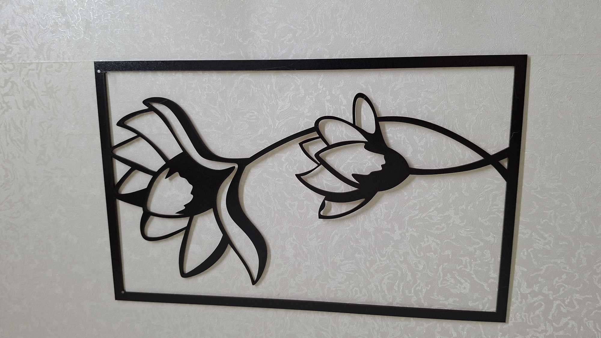 The image displays a black metal wall art piece mounted on a light-colored, textured wall, likely in an indoor setting in Большой Букор, Russia. The artwork is a rectangular panel featuring a stylized floral design, with two prominent flowers connected by a flowing stem. These elements are cut out from the black metal, creating a striking silhouette effect against the subtle, marbled or swirled pattern of the off-white wall. The thin black frame of the art piece includes small holes in the corners, suggesting it is affixed to the wall with fasteners. The lighting is even and soft, indicative of an interior space. There are no people, activities, specific time of day, weather conditions, or visible text present in the image.