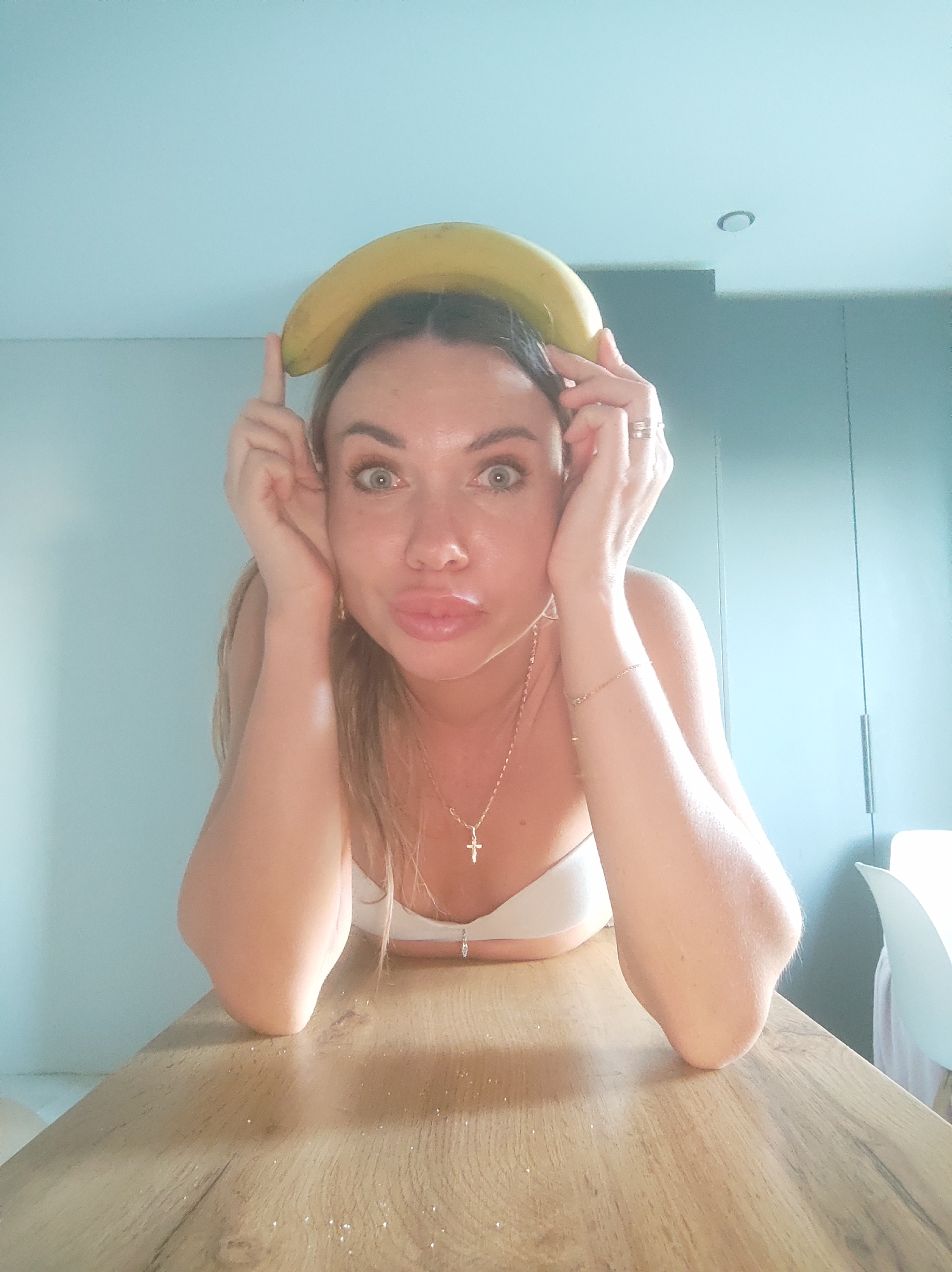 A woman poses with a banana balanced on her head. She rests her arms on a wooden table and looks directly at the camera with a playful expression. She wears a white top and gold jewelry, including a necklace with a cross pendant and a bracelet. The background includes a light blue wall. The setting appears to be indoors. The location is not discernible from the image.