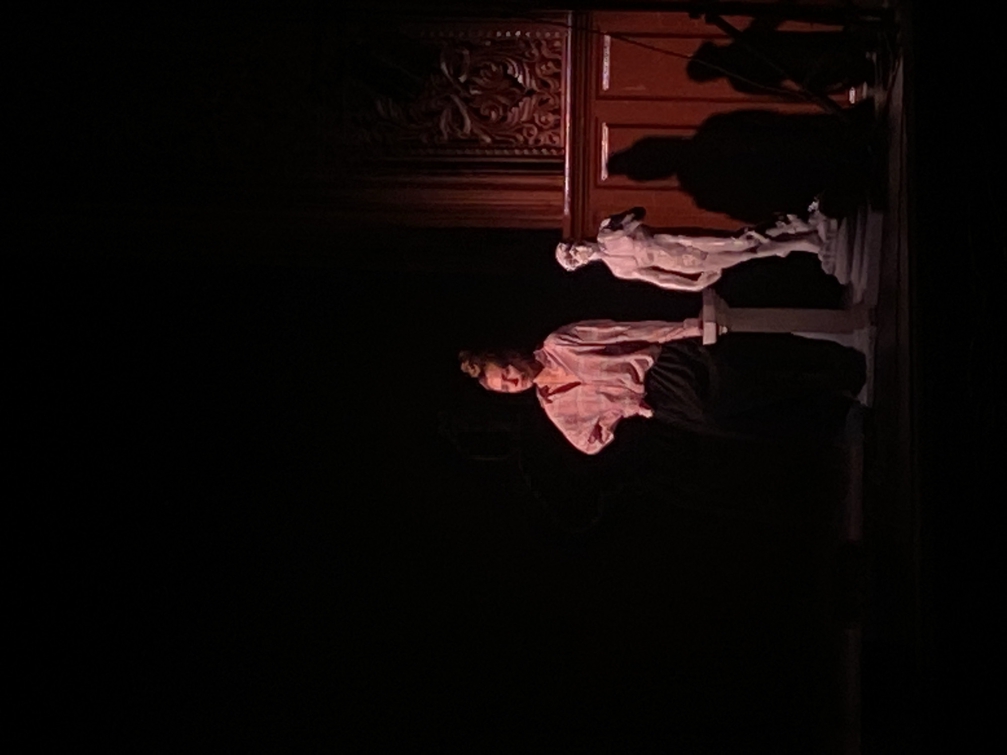 The image depicts a dimly lit stage setting with a woman wearing a pink patterned shirt and dark skirt standing next to a white sculpture. Behind them is an ornately carved wooden structure, possibly a backdrop or set piece. Above the sculpture, shadows suggest lighting equipment. The location, Rusanovo, Russia, is mentioned without any elements to verify it.
