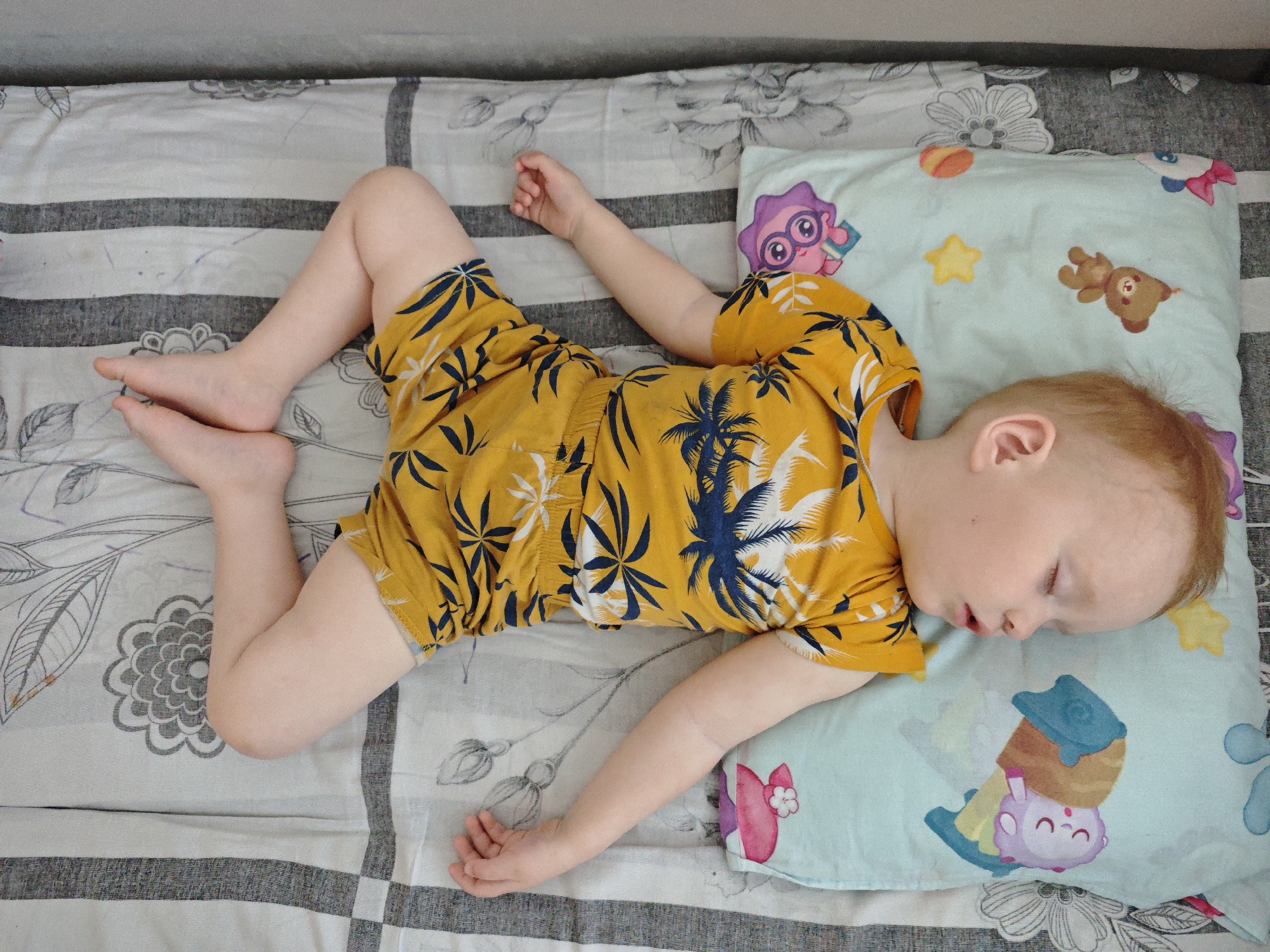 A fair-skinned child wearing a yellow shirt and shorts patterned with blue palm trees is asleep on a bed. The bed has a gray and white striped and floral patterned sheet. The child's head rests on a light blue pillow printed with cartoon characters and stars. The location of Markovskii, Russia, is not discernible from the image.