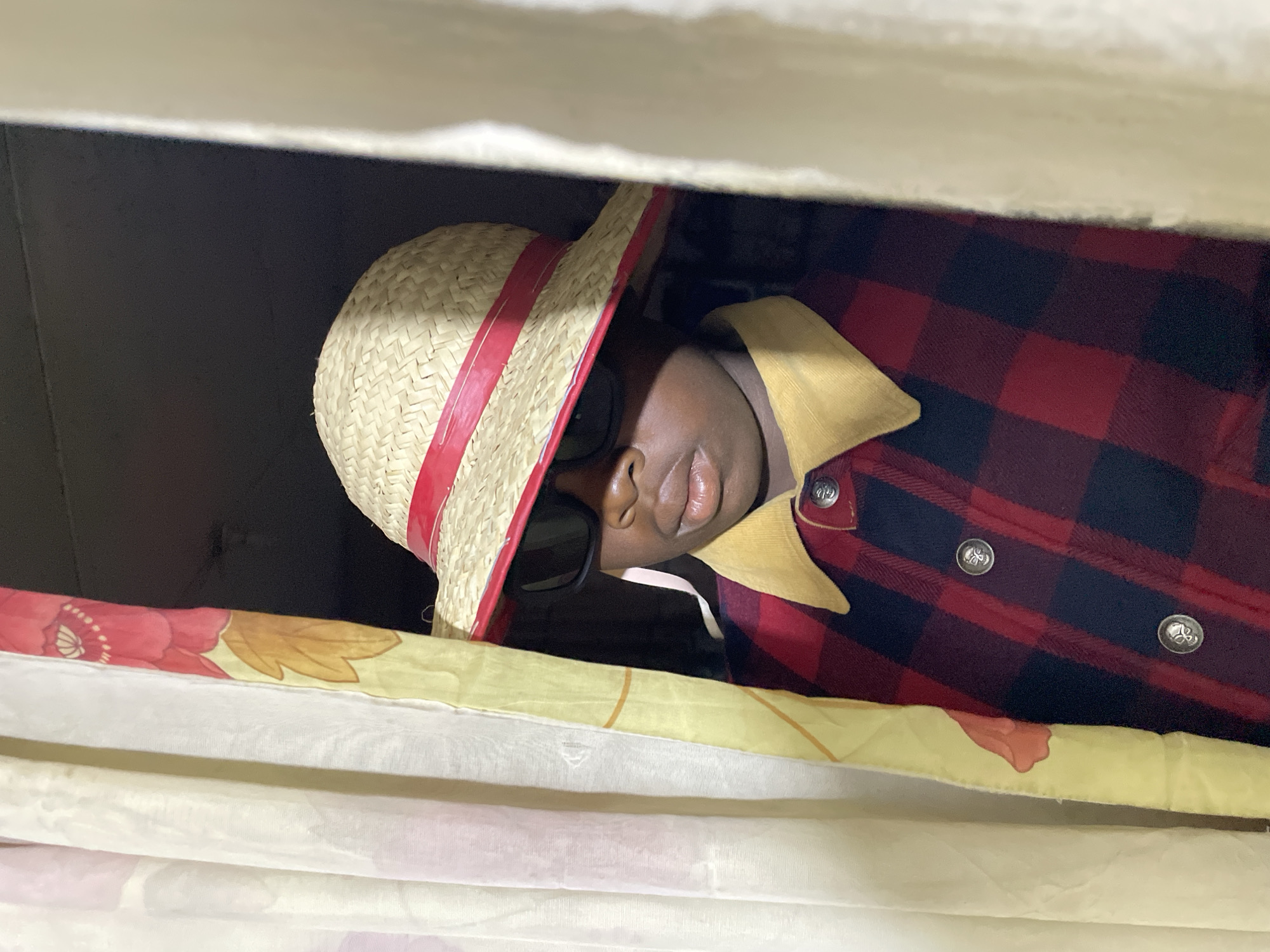 A person is partially visible, peeking from between two layers of fabric. The individual has dark skin and is wearing a straw boater hat with a prominent red band, paired with dark sunglasses that obscure their eyes. Their nose and mouth, featuring full lips, are clearly visible. They are dressed in a red and black plaid flannel shirt or jacket, characterized by a light brown or tan corduroy collar and metallic silver buttons.
The person is framed by two distinct fabric elements. Above, there is a plain, light-colored fabric, while below, a vibrant yellow fabric adorned with floral patterns, including red and golden-brown designs, is visible. The background behind the person is dark and indistinct, suggesting they are situated in a shadowed area or looking out from a confined space. No text is visible in the image. The scene is set in Jos, Nigeria.