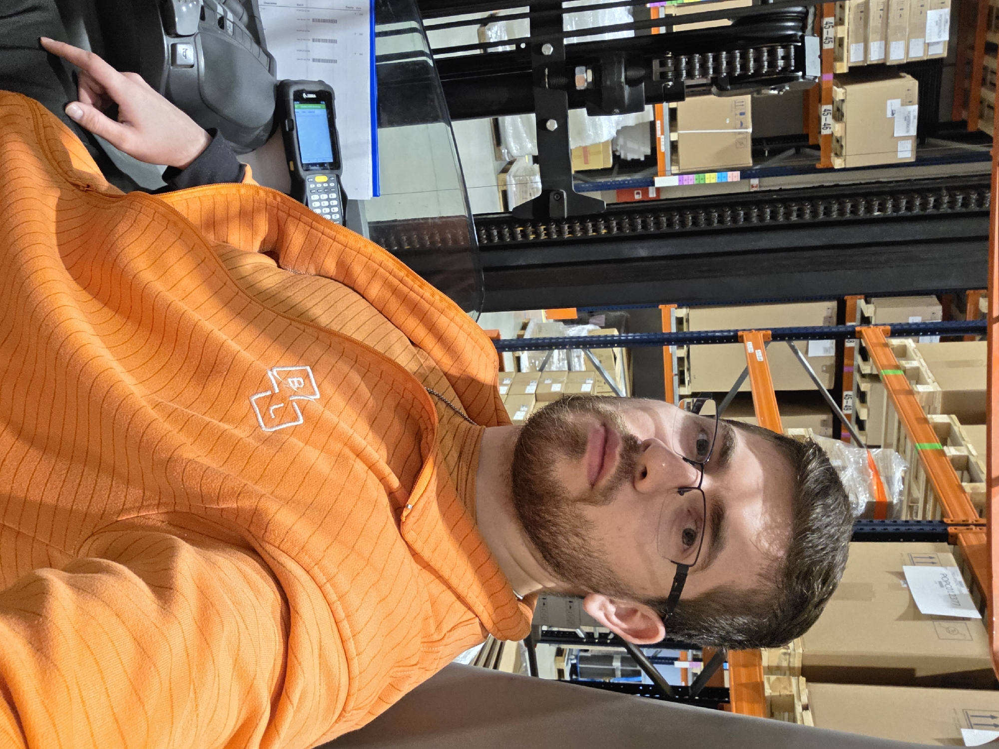 A man with glasses and a beard wearing an orange hoodie sits in the driver's seat of a forklift inside a warehouse, potentially in Plovdiv, Bulgaria. Shelves with boxes are visible in the background. A handheld scanner and paperwork are beside him. He is looking at the camera.
