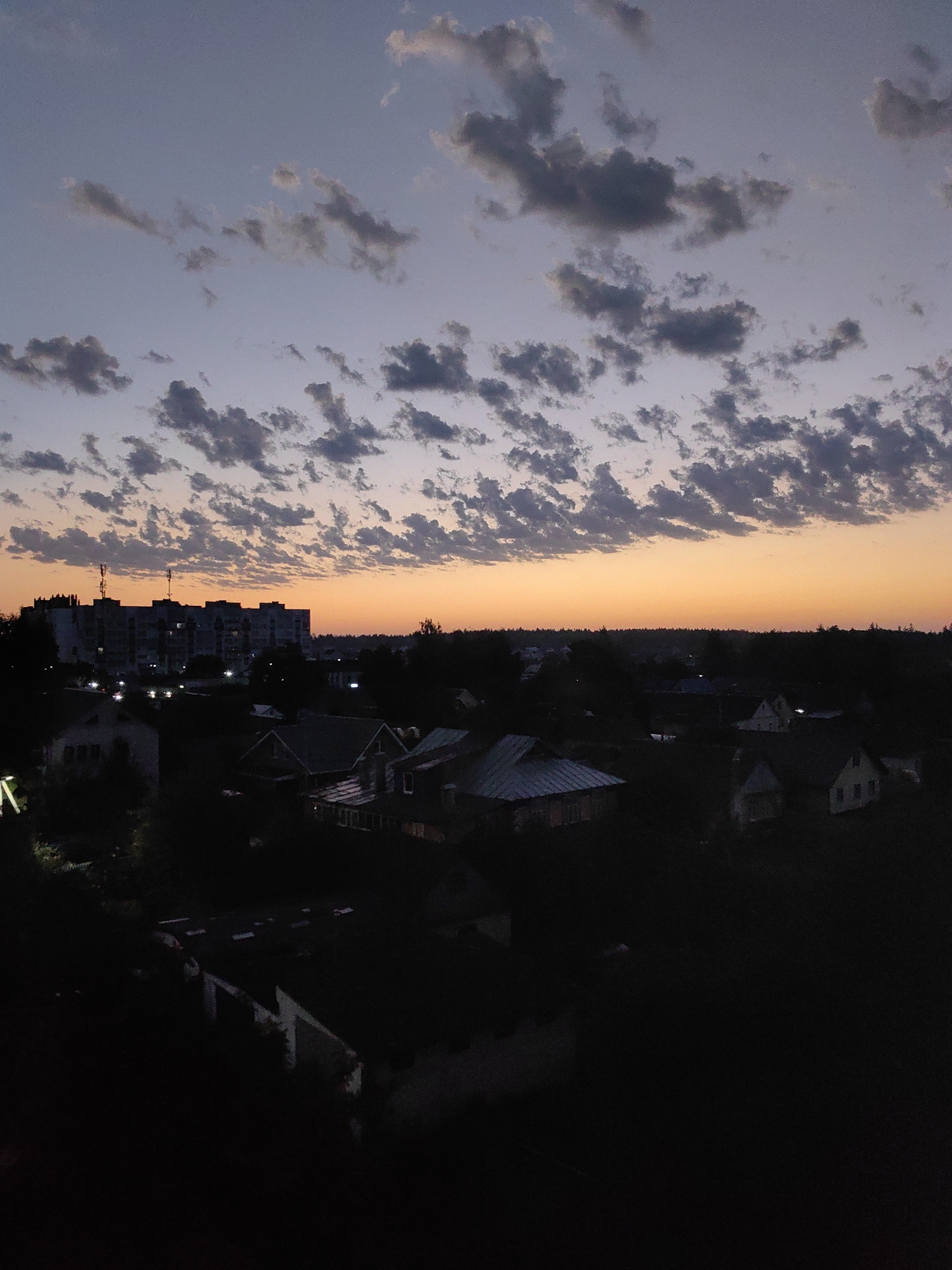 This is a wide-angle, aerial photograph taken at dusk over the city of Fanipal, Belarus. The sky is a gradient of deep blue at the top to a warm orange glow near the horizon, indicating the setting sun. Scattered across the sky are numerous small, dark clouds, creating a textured effect.
In the mid-ground, a cluster of buildings is visible. The most prominent structure is a multi-story apartment building with visible windows and at least two communication towers on its roof. To the right of the apartment building, several smaller residential houses with pitched roofs are discernible. The lower portion of the image is dominated by dark shadows, obscuring most of the foreground and many of the details of the houses. Some scattered lights are visible within some of the buildings, suggesting indoor illumination. Trees and foliage are present throughout the scene, particularly in the mid-ground and along the horizon, adding to the overall dimness of the lower half of the photograph. The scene appears to be residential, with a mix of apartment blocks and smaller homes. There are no visible people in the image.