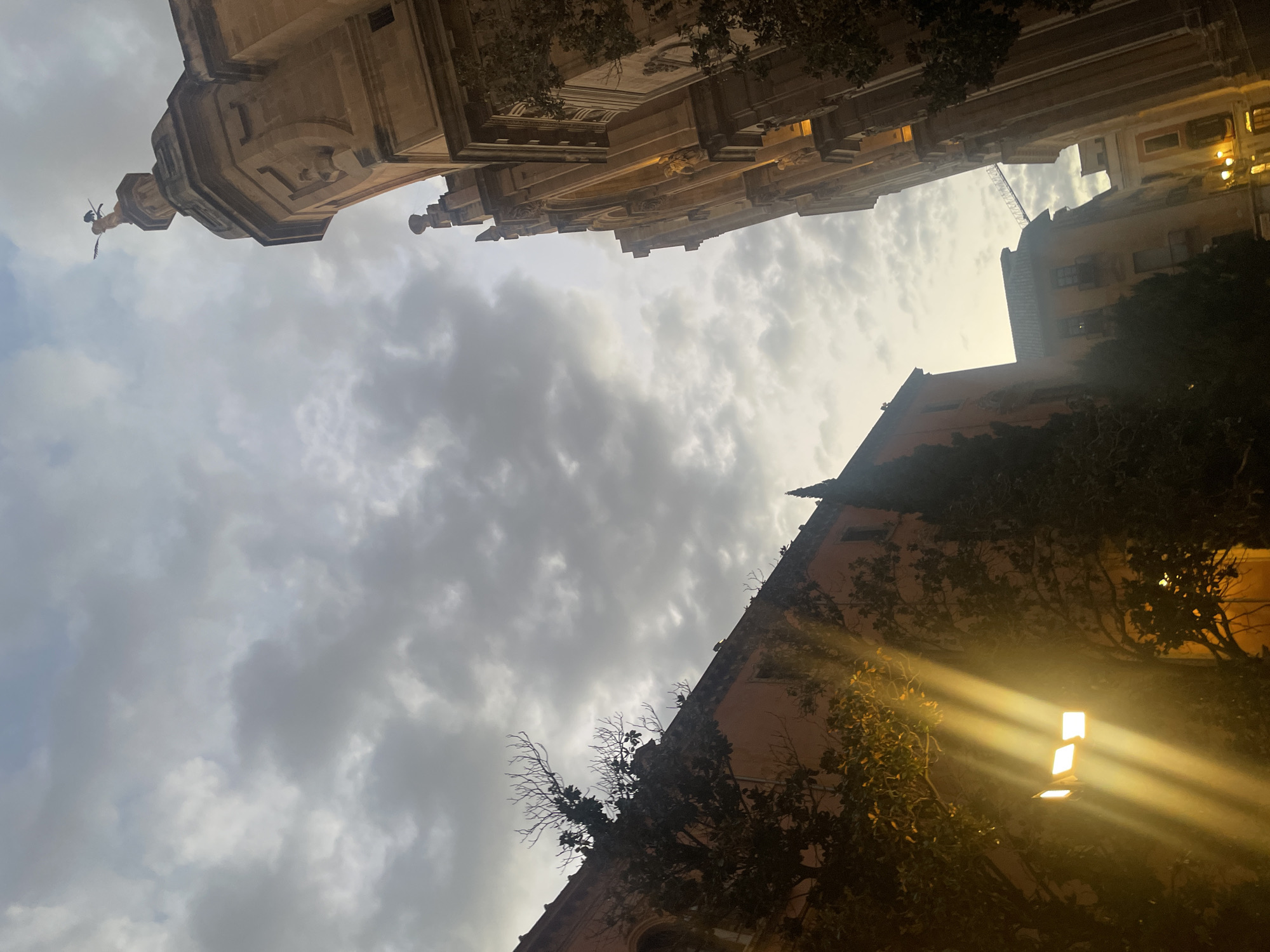 This is an upward-facing, low-angle photograph taken in Granada, Spain, capturing the interplay of a dramatic sky with architectural elements and trees. The sky is filled with heavy, textured clouds, with patches of lighter sky peeking through.
On the right side of the frame, a tall, ornate building, likely a church or cathedral, dominates the view. Its weathered stone facade is intricately detailed with carvings and architectural features, suggesting historical significance. A small, dark figure, possibly a statue or a bird, is perched atop the building's spire, silhouetted against the cloudy sky.
To the left, a lower, orange-hued building stretches across the frame. Trees with bare branches and dense foliage are seen in front of and on top of this structure. A bright, modern light fixture is positioned low on the left, emitting strong, golden beams of light that cut through the scene, illuminating the trees and casting dramatic rays across the building.
In the background, other buildings of varying heights and architectural styles are visible, hinting at a dense urban environment. The overall impression is one of historical grandeur set against a moody, atmospheric sky, with the unexpected modern element of the bright spotlight adding a contemporary contrast. The lighting suggests it is either dawn or dusk.