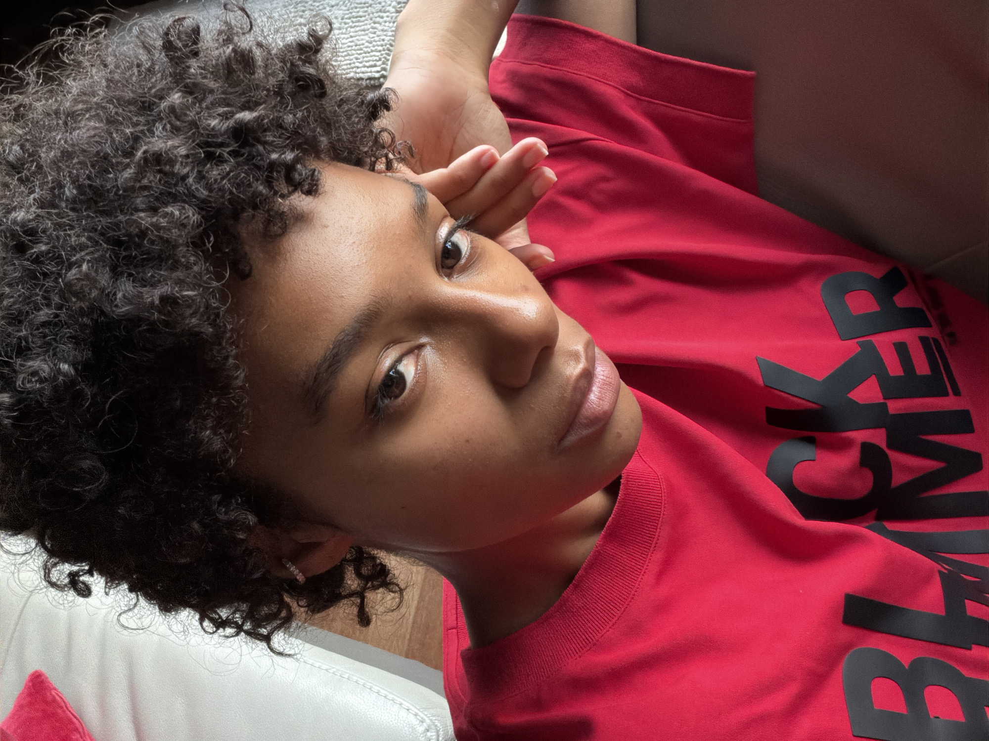 The image shows a close-up, high-angle shot of a young Black person with curly dark hair, wearing a bright red t-shirt with large black lettering that appears to say "BLACK LIVES MATTER." The person is leaning their head on their hand, with their face turned towards the camera. Their skin is a rich brown, and they have dark eyes and full lips. A faint sparkle is visible on their earlobe, suggesting they might be wearing an earring. The background is softly blurred, showing what appears to be the edge of a light-colored sofa or chair. The lighting suggests an indoor setting, possibly during the daytime. The scene is a portrait of the individual, with a focus on their facial features and the message on their shirt. There is no indication of the city of Champs-sur-Marne, France, in the image itself.