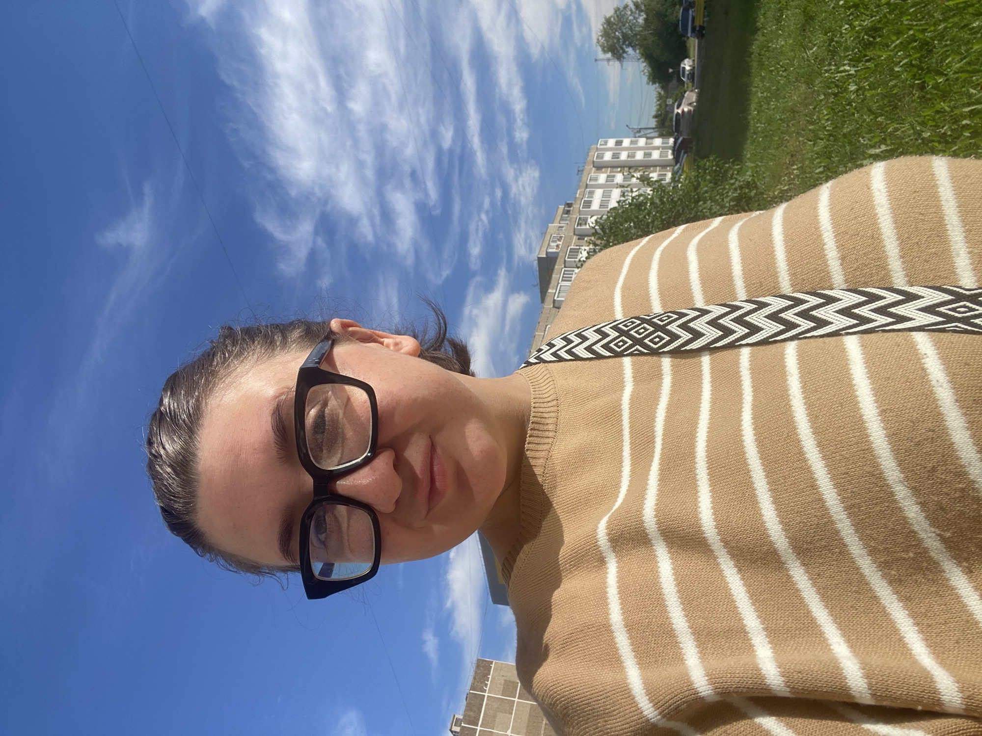 A woman wearing black-rimmed glasses and a tan and white horizontally striped sweater stands outdoors. She has her hair pulled back and is looking at the camera. A patterned strap, likely from a bag, is visible across her chest. The background shows a bright blue sky with wispy white clouds, a multi-story building, trees, and a grassy area with a road and parked cars. The scene appears to be in a residential area in the city of Chaikovsky, Russia.