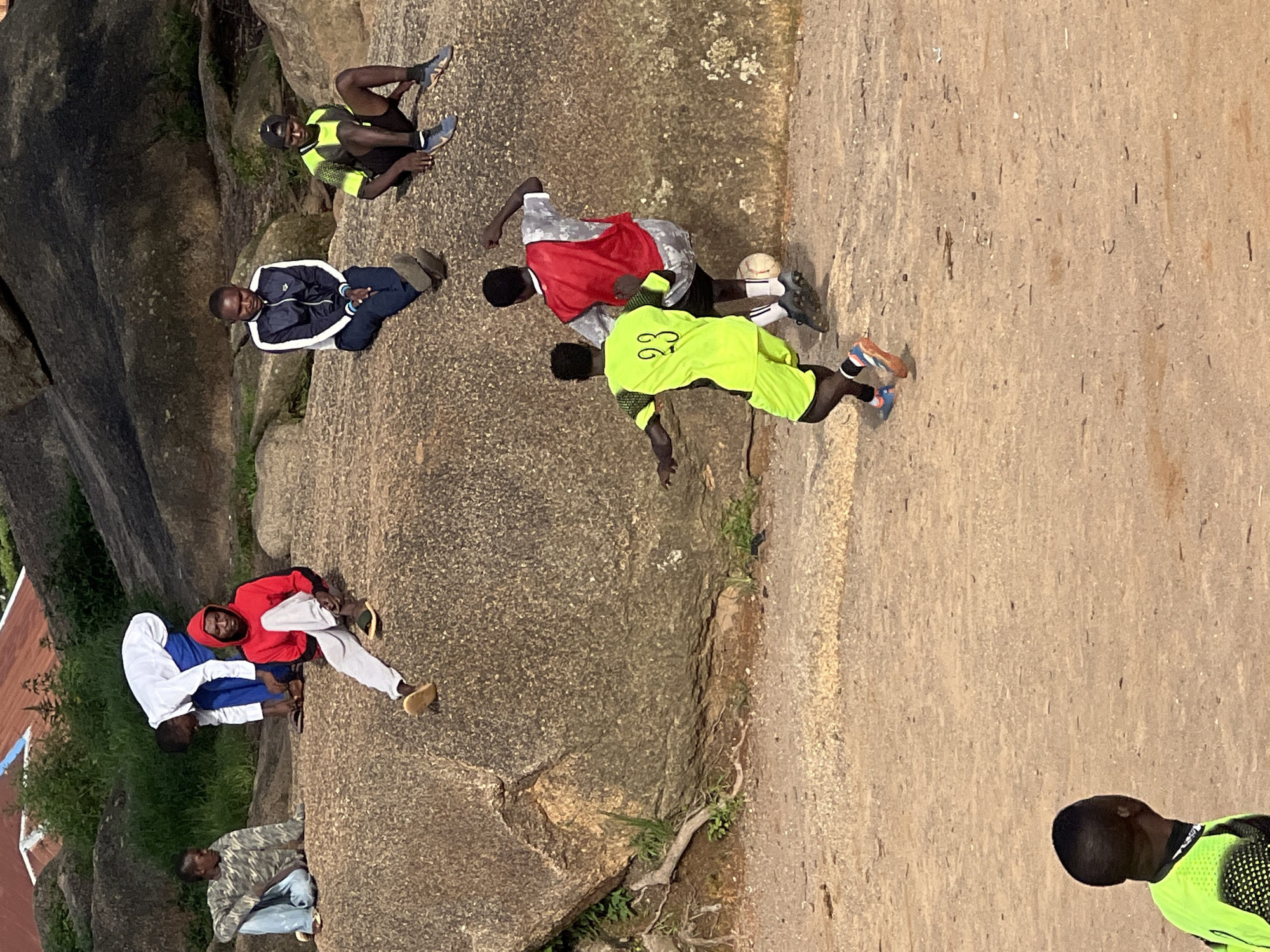 A group of young men are gathered on and around a large rock formation. In the foreground, two men are playing soccer on a dirt field. One man, wearing a bright yellow jersey with the number 23, is kicking the ball. The other man, wearing a red jersey, is positioned to block the shot. Several other men are sitting on the rock formation in the background, observing the game or resting. One man in a red hoodie and grey pants is seated on the rock, looking towards the soccer players. Another man in a camouflage jacket and blue jeans is also seated on the rocks to the left. Further back, two men are seated on the rocks; one in a dark jacket and white shirt, and another in a neon yellow and black soccer jersey and black shorts. The environment appears to be semi-arid, with sparse vegetation and large rock outcrops, consistent with the landscape found in Jos, Nigeria. The lighting suggests it is daytime, likely late afternoon, given the shadows.