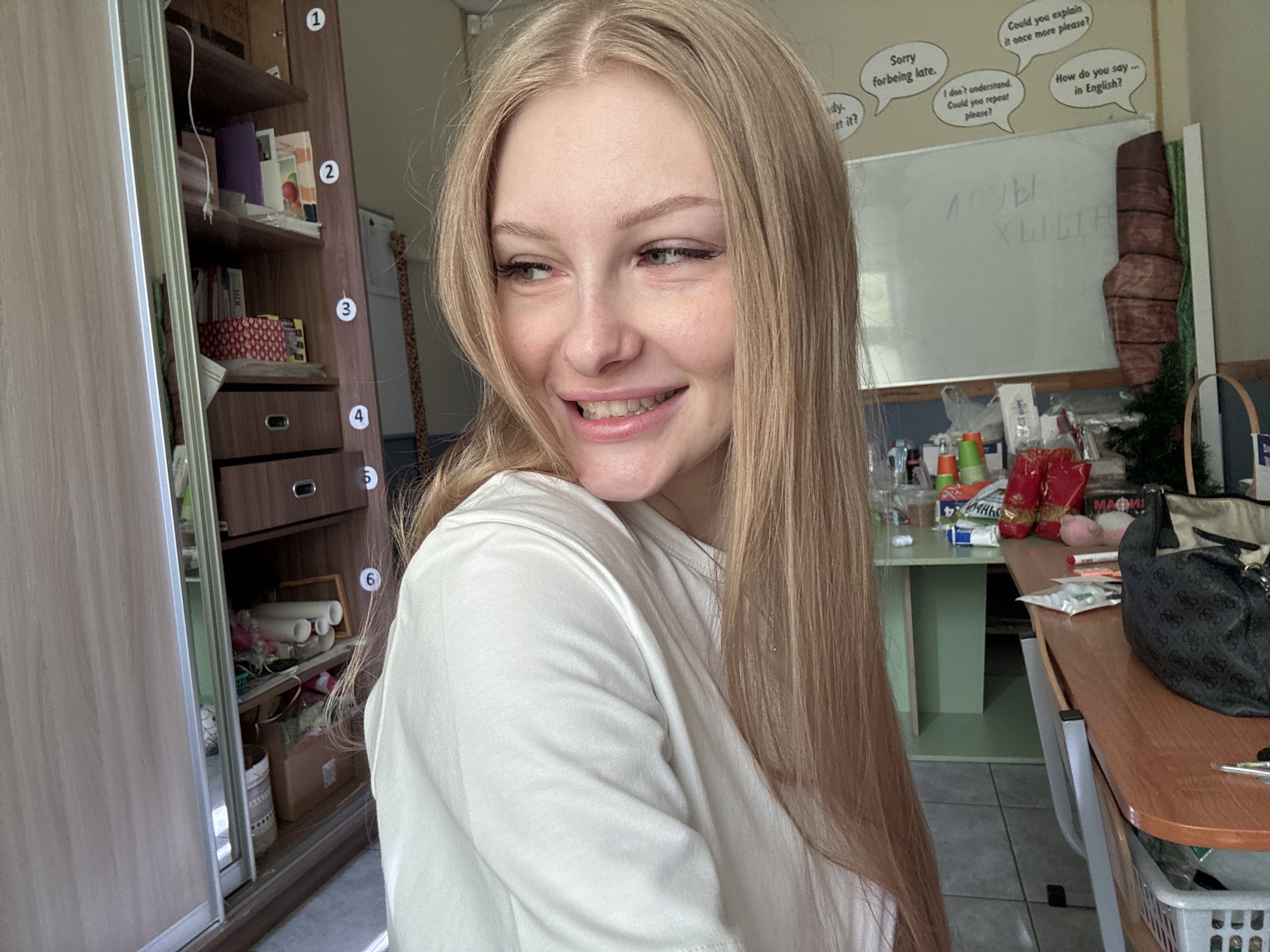 A young woman with long blonde hair and fair skin is pictured from the chest up, looking to her left with a slight smile. She is wearing a white t-shirt. The background includes a bookshelf on the left, a whiteboard with writing in Cyrillic script and speech bubbles containing English phrases on the back wall, and a wooden desk in the foreground on the right. The overall setting appears to be a classroom or study area. The specific location in the city of Perm, Russia is not discernible from the image.