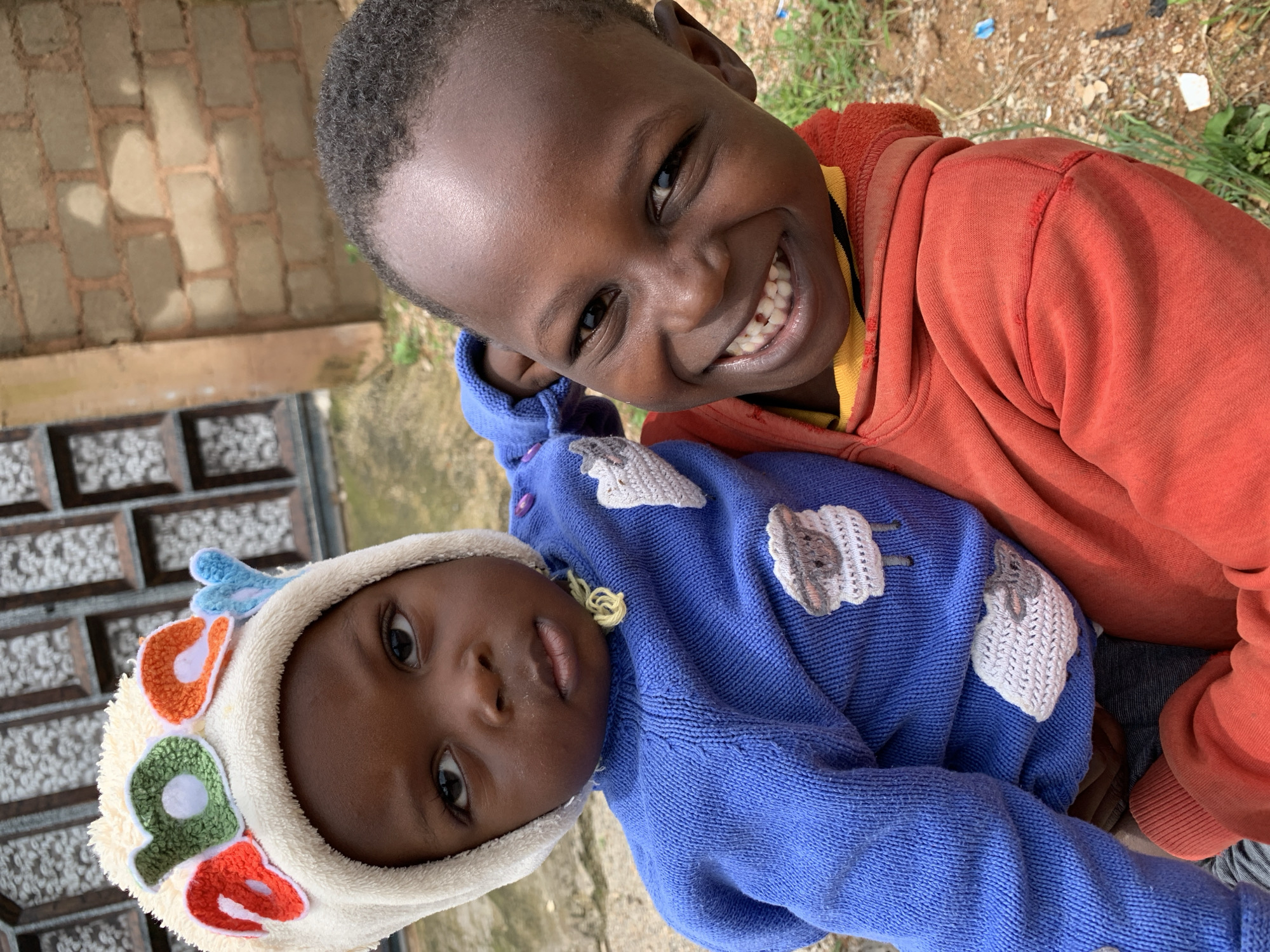 Two young Black children are positioned in a close-up shot, outdoors. The child in the foreground, wearing a red hooded sweatshirt and a yellow shirt underneath, grins widely, revealing a gap in their front teeth. The child's left arm is bent, holding the other child. The second child, positioned behind and slightly to the left, wears a blue knitted sweater and a cream-colored hat decorated with colorful letters. This child looks directly at the camera with a more serious expression. The background consists of a brick wall and a partially visible window with a decorative lattice. The ground is covered with dirt and sparse green grass. The overall impression is a candid, warm interaction between the two siblings.