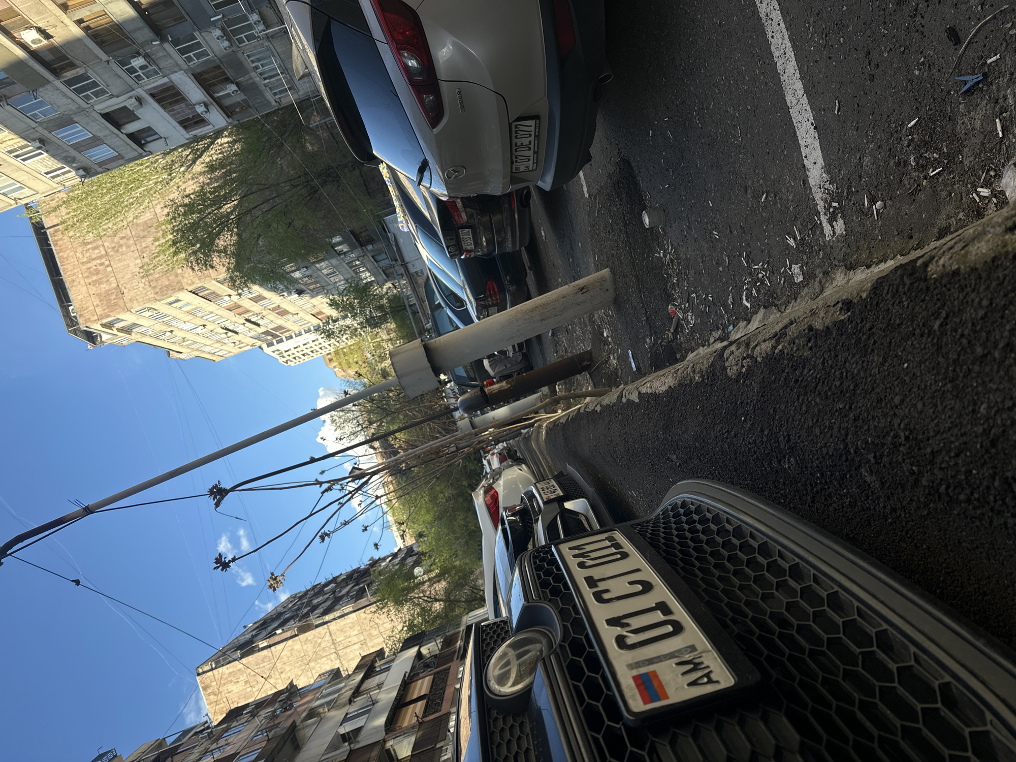 The image captures an outdoor urban street scene from a low-angle perspective, with the original photo rotated 90 degrees clockwise, making the ground appear on the right side and the sky on the left.
In the foreground, a dark car partially obstructs the view, showcasing its hexagonal grille and front license plate. The plate, "01 CT 030", clearly displays the Armenian flag and the country code "AM", indicating the location is Armenia. Behind it, a white Mazda SUV is parked, its rear visible with the license plate "07 DE 077". Further down the street, another car has the license plate "84 SS 092". All cars are parked parallel to a concrete curb.
The asphalt road features a white line, commonly used for parking or lane demarcation. The area next to the curb is littered with numerous cigarette butts, small pieces of paper, plastic, and a blue clothes peg. A white, weathered barrier or pole is positioned in the immediate foreground, separating the viewer's perspective from the parked vehicles.
Multi-story buildings with many windows and external air conditioning units line the street. Their architecture suggests an older, possibly Soviet-era urban setting. Trees with fresh green leaves are visible between the buildings, indicating spring or early summer. The sky is bright blue with a few faint clouds, and several utility wires or cables crisscross the upper portion of the frame.
The scene depicts a typical daylight moment in an Armenian city, with clear weather and no discernible human activity.