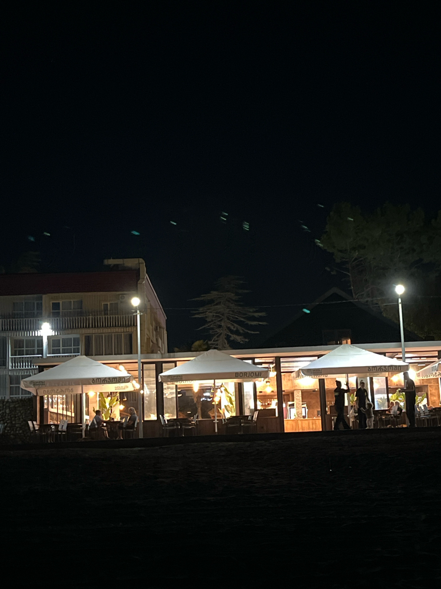 This is a nighttime photograph of a beachfront restaurant in Ureki, Georgia. The restaurant features a covered outdoor seating area with white umbrellas and illuminated interior dining spaces. Several people are visible, including patrons seated at tables and staff members serving or walking around. The building behind the restaurant appears to be a multi-story hotel or guesthouse. The foreground consists of dark sand, suggesting the restaurant is situated on a beach. The sky is dark, with some faint streaks of light visible, possibly from streetlights or distant vehicles. The overall impression is of a lively evening scene at a seaside establishment.