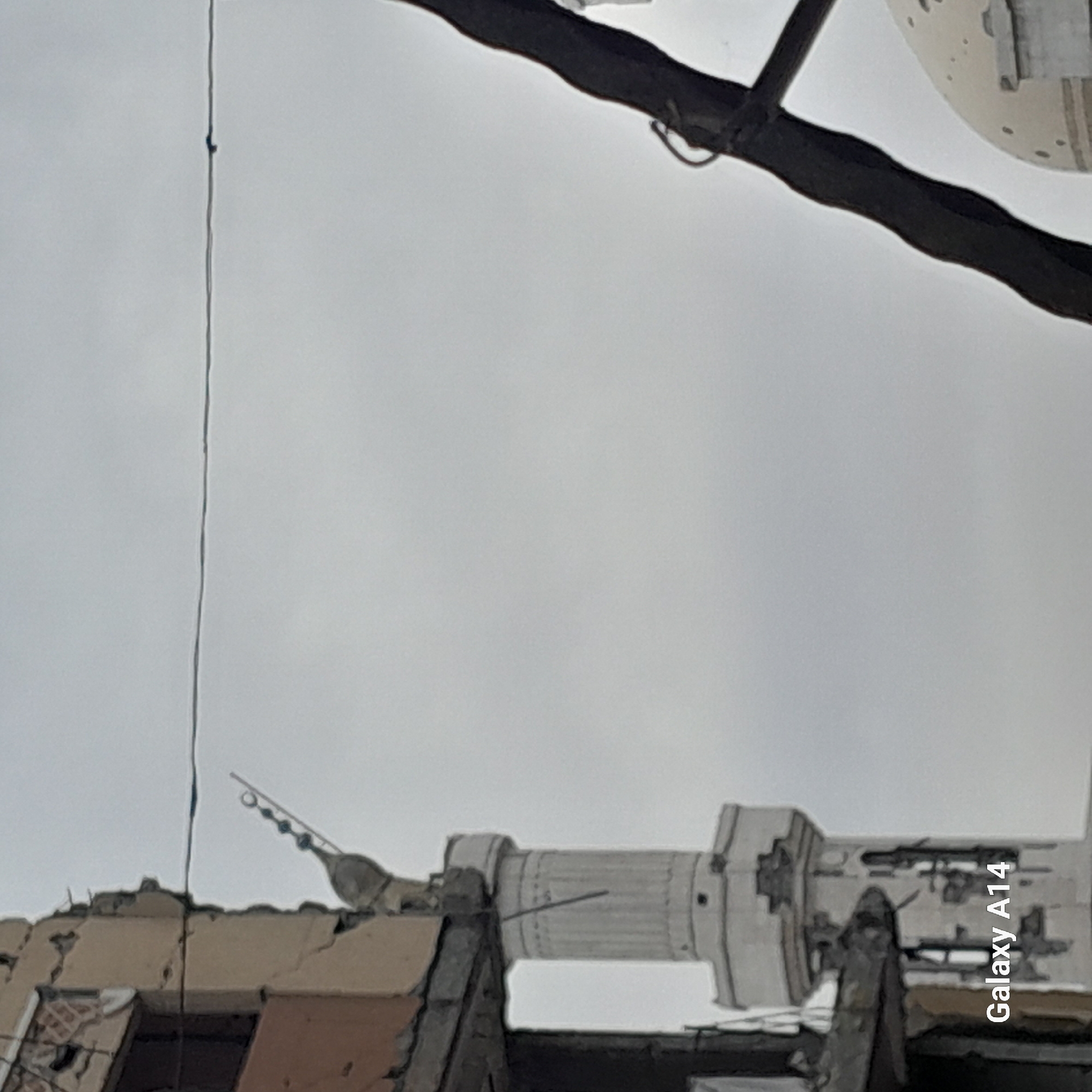 The image shows a minaret of a mosque in what appears to be a damaged urban environment. The minaret is white with cylindrical sections. The foreground consists of rubble and damaged buildings. Overhead, there are electrical wires. The sky is overcast. The photo was taken with a Galaxy A14 phone. The location is unknown.
