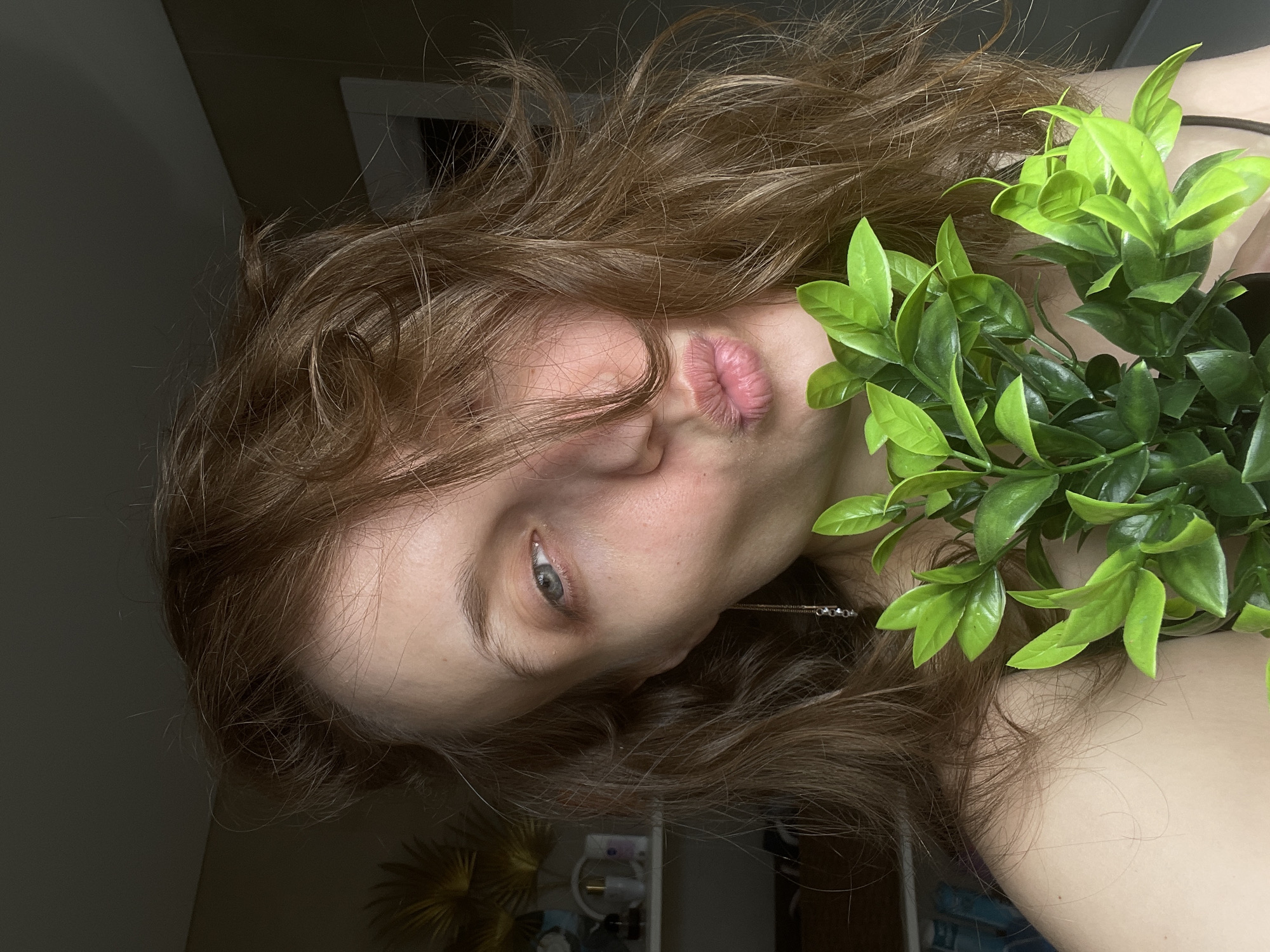 A woman is lying on her side with green foliage partially obscuring her neck. The woman has auburn hair and is looking directly at the camera with one eye open. Her lips are pursed. The background is a dark room with a doorway visible in the upper left. A portion of a dresser and a decorative plant are visible in the lower portion of the image. The location is not identifiable as Podshivalovo, Russia based solely on the provided image.